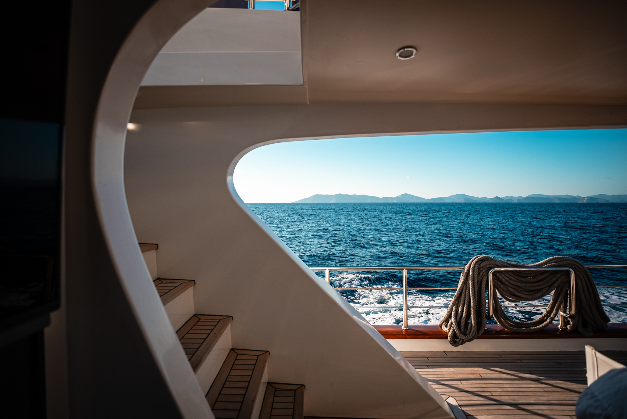 View from inside a yacht looking out to the ocean, with curved stairs on the left, thick ropes coiled on the deck railing, and distant mountains visible across the blue water under a clear sky.
