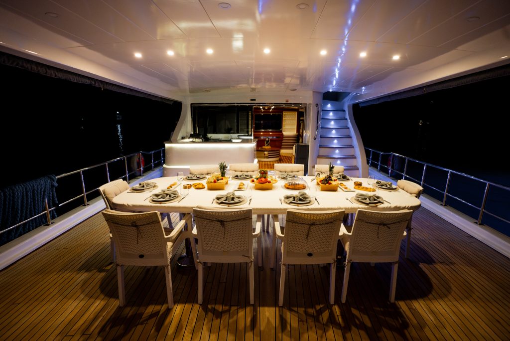 A long dining table set with plates, glasses, and food is arranged on the deck of a yacht at night, with white chairs and soft lighting creating an elegant, inviting atmosphere.