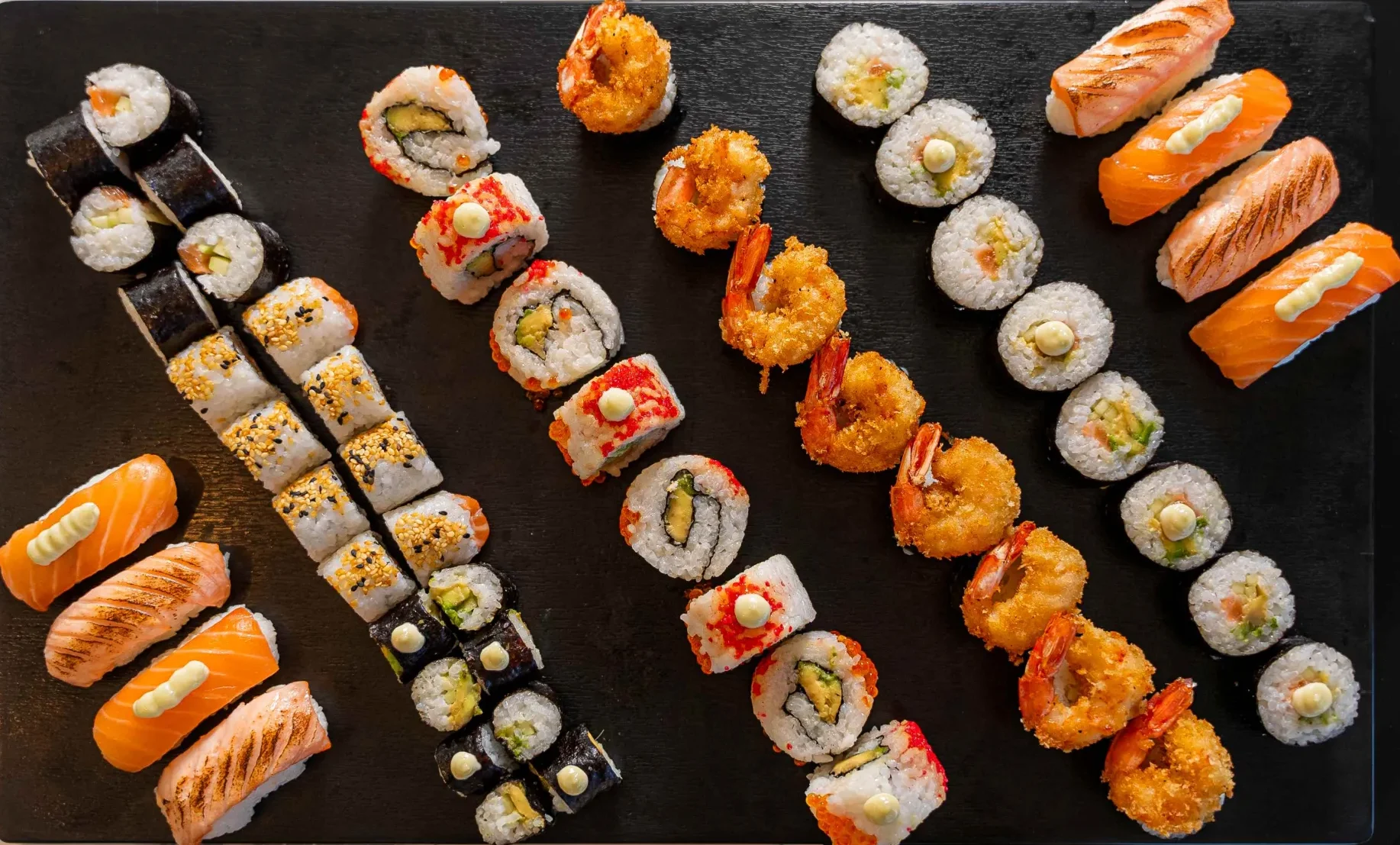 A rectangular black plate displays assorted sushi, including nigiri, maki rolls, and tempura shrimp rolls, arranged in neat parallel rows on a marble surface.