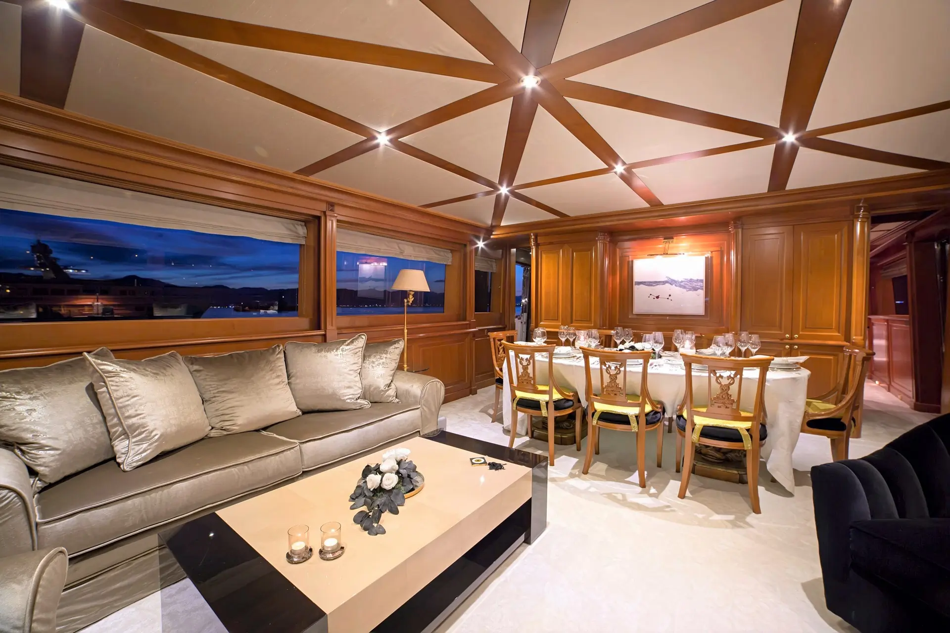 Elegant yacht interior with a beige sofa, a coffee table, and a formal dining area set for eight. Warm wood paneling, large window with a night view, and stylish ceiling lighting create a luxurious atmosphere.