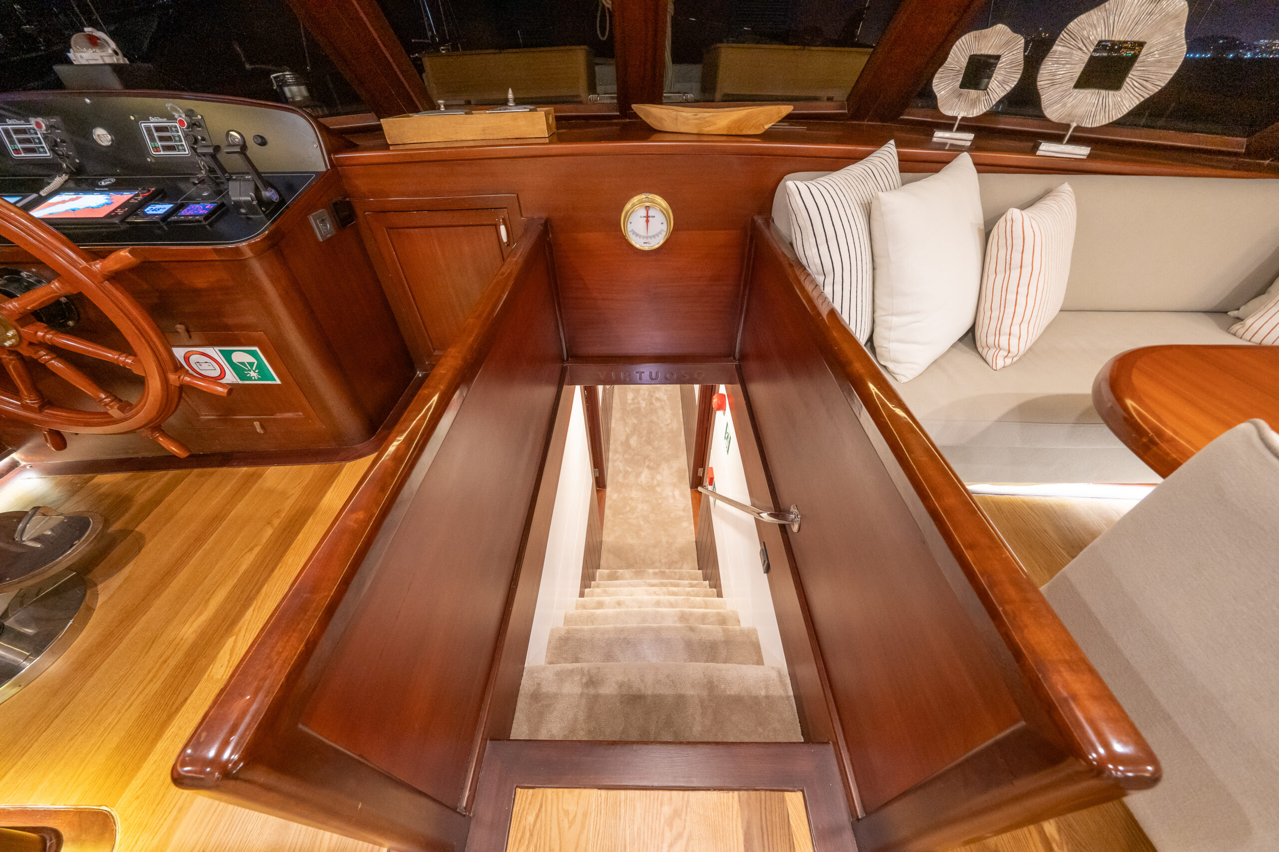 VIRTUOSO A wooden staircase with carpeted steps leads down from a yachts main cabin, surrounded by polished wood paneling. The helm and seating area with cushions are visible beside the stairway.