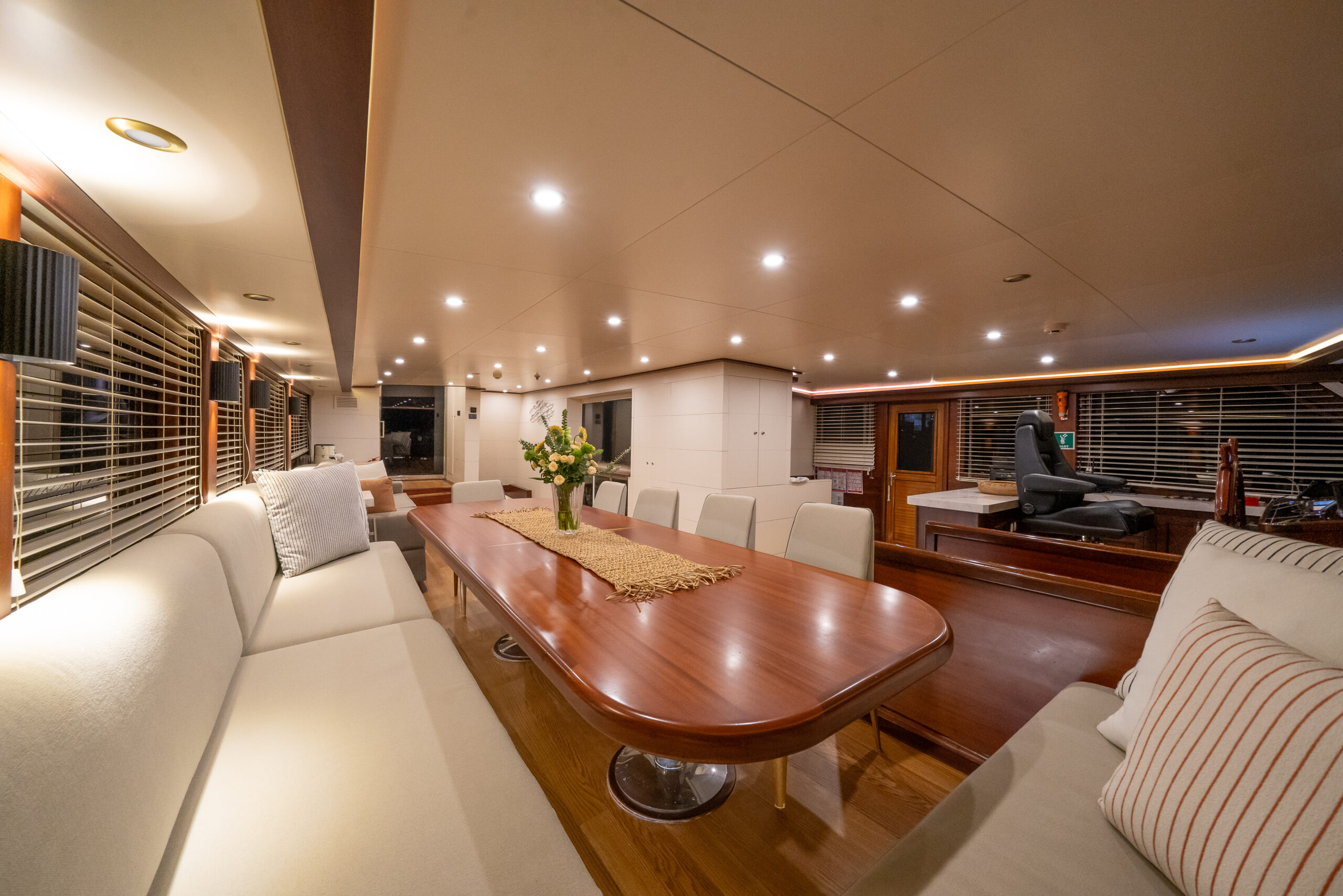 VIRTUOSO A spacious, well-lit yacht interior features a long wooden dining table with white cushioned seats, decorative pillows, a flower arrangement, and large windows with blinds, creating a modern and inviting atmosphere.