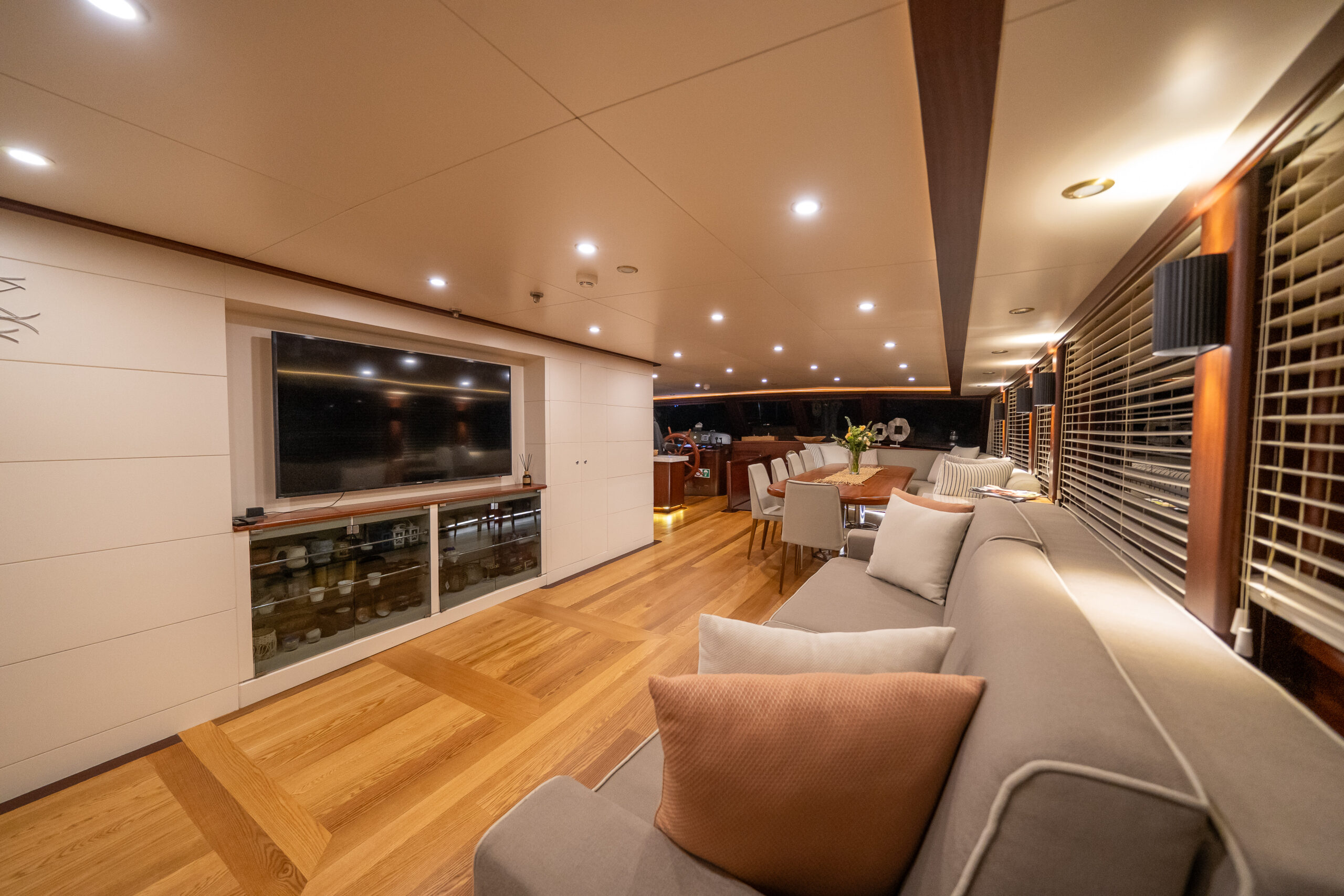 VIRTUOSO A spacious, modern yacht interior with wood floors, neutral sofas with cushions, a large flat-screen TV, dining table, and large windows with blinds, warmly lit by ceiling lights.