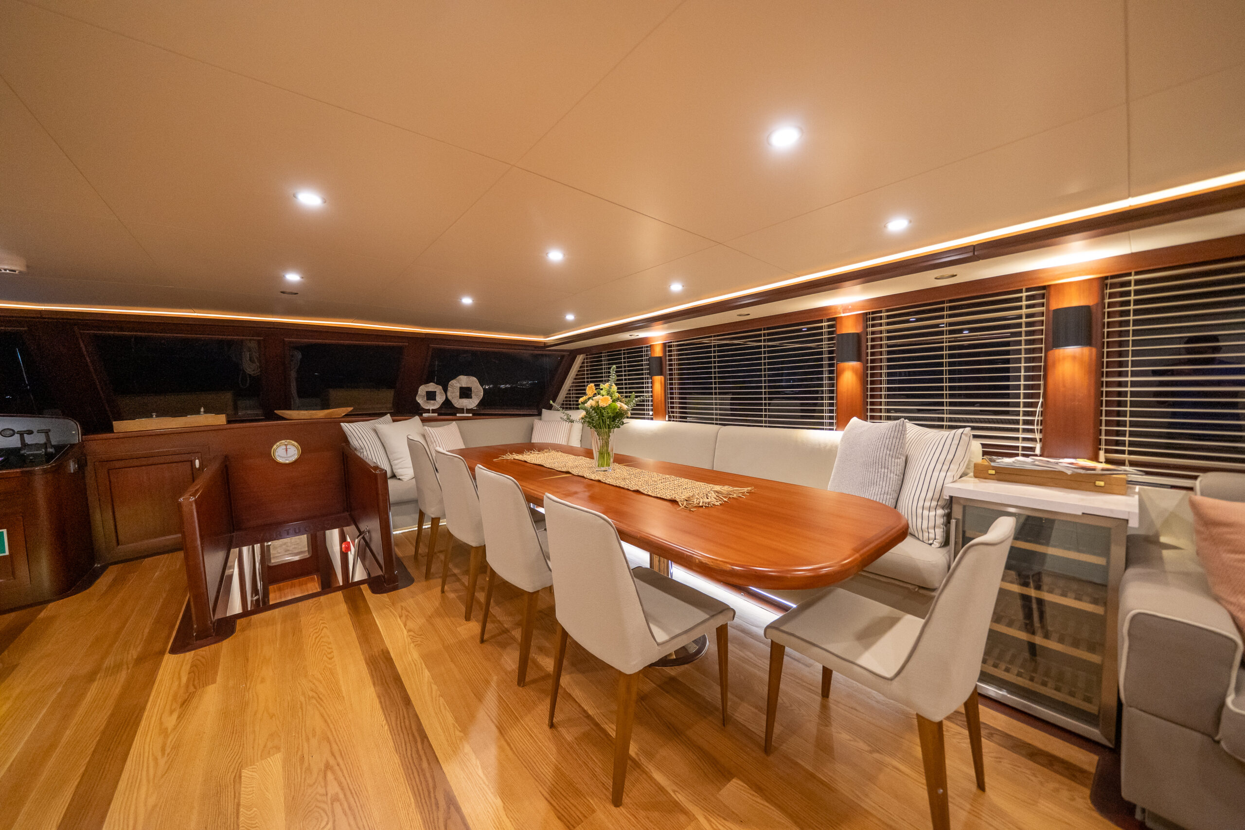 VIRTUOSO A luxurious yacht interior with a polished wooden dining table, cushioned chairs, and benches. The space is warmly lit, has wooden floors, large windows with blinds, and a small bar fridge and flower centerpiece on the table.