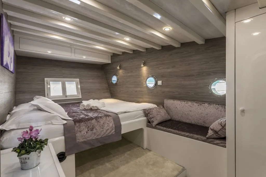 A cozy yacht cabin with a double bed, rolled towels, small round windows, a cushioned bench, modern lighting, and a small potted plant on a white surface. The decor is sleek with wood-paneled walls and soft tones.