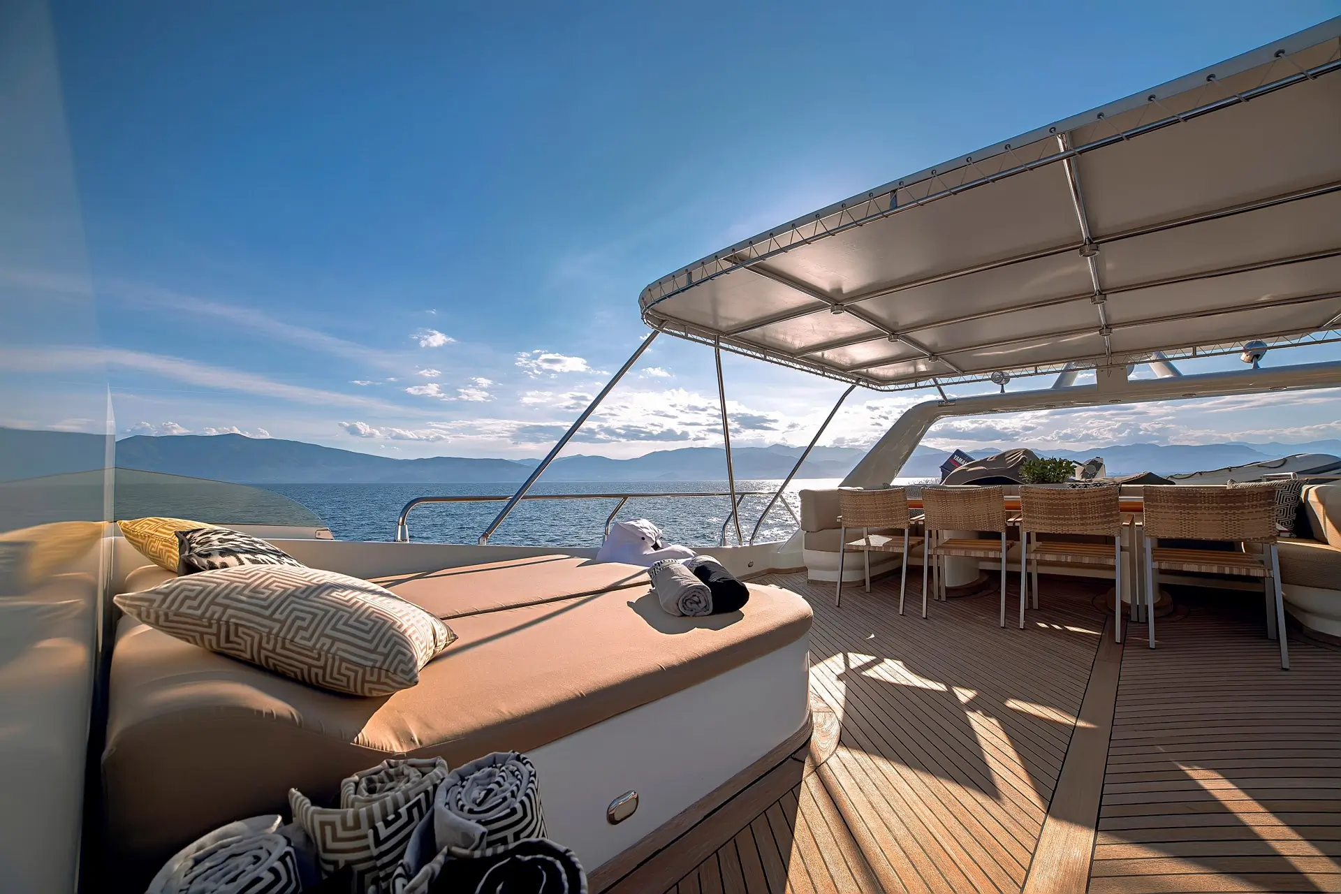 Luxurious yacht deck with cushioned lounge chairs, rolled towels, and a shaded dining area, overlooking calm blue sea and distant mountains under a bright, sunny sky.