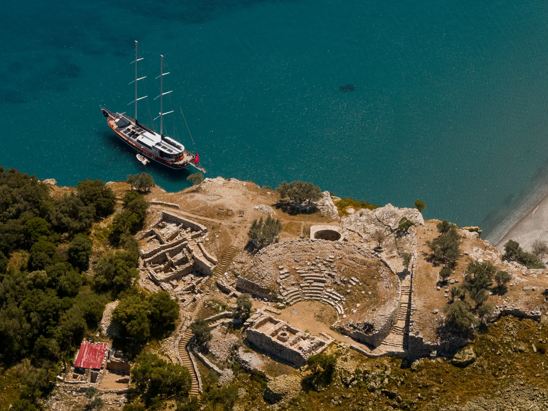 Aerial view of ancient stone ruins, including a small amphitheater, near the turquoise sea with a docked sailboat. Dense green trees surround the site, and a red-roofed building is visible among the ruins.