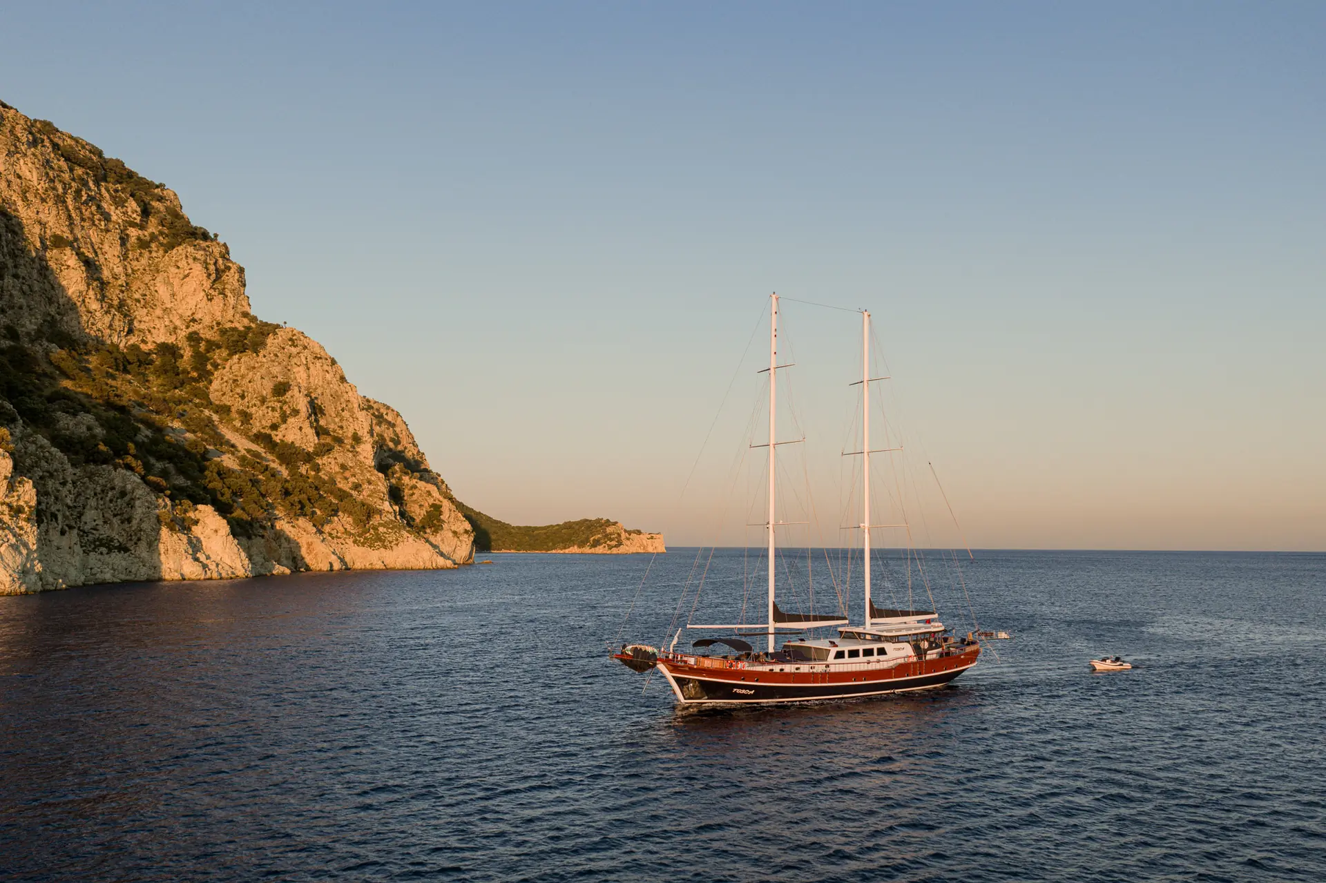 A large sailboat with two masts floats on calm blue water near rocky cliffs at sunset, with a small motorboat nearby and warm sunlight illuminating the scene.