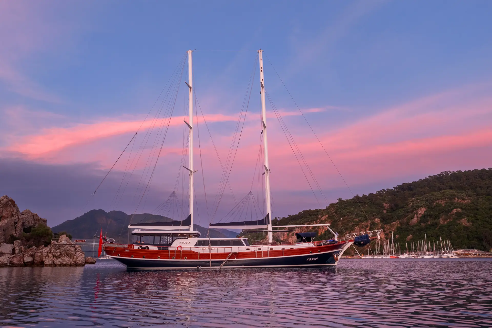 A large sailboat with two masts floats on calm water at sunset, with pink clouds in the sky and green hills in the background.