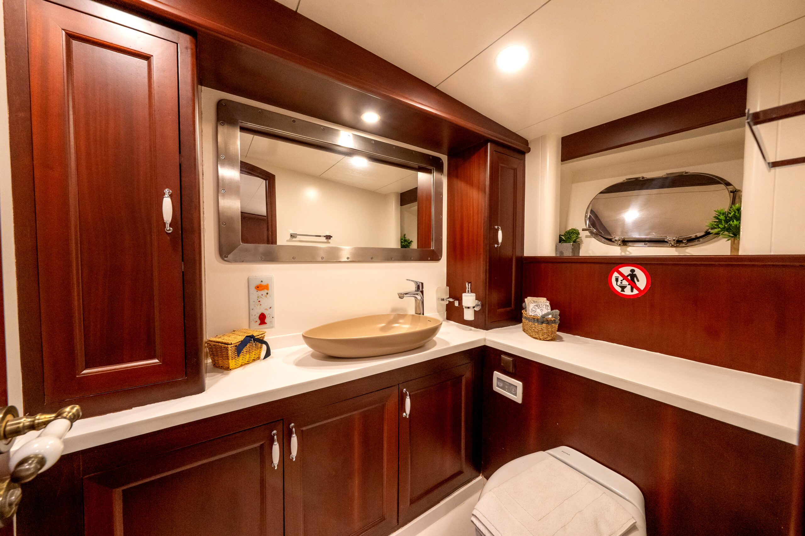 VIRTUOSO A yacht bathroom with wooden cabinets, a modern vessel sink, a large mirror, porthole window, and signs indicating “no toilet paper” in the toilet. The space is well-lit with simple decor and toiletries on the counter.