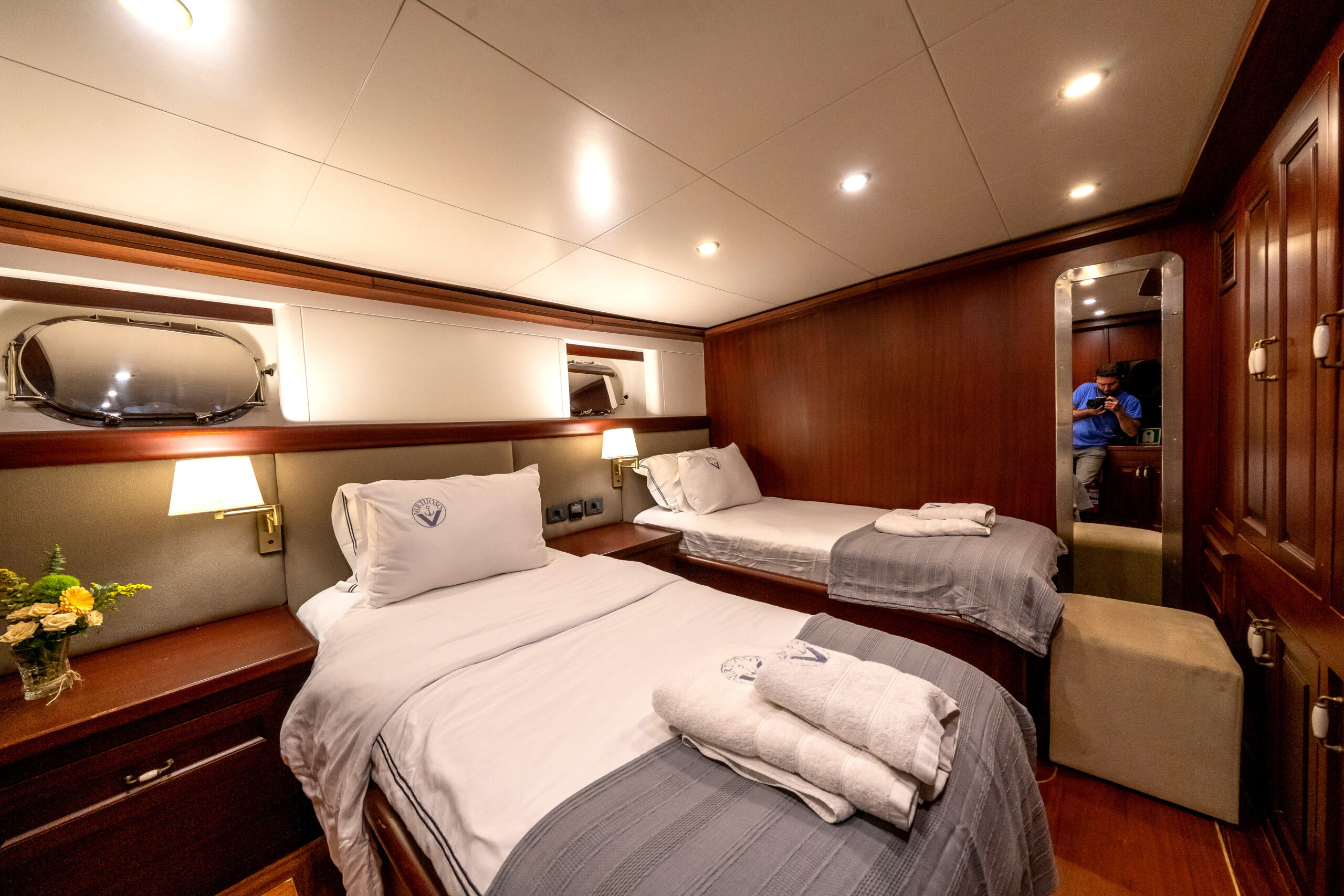 VIRTUOSO A cozy yacht cabin with two beds, wood-paneled walls, white linens, a porthole window, bedside lamps, and a bouquet of flowers; a person is reflected in the mirror taking a photo.