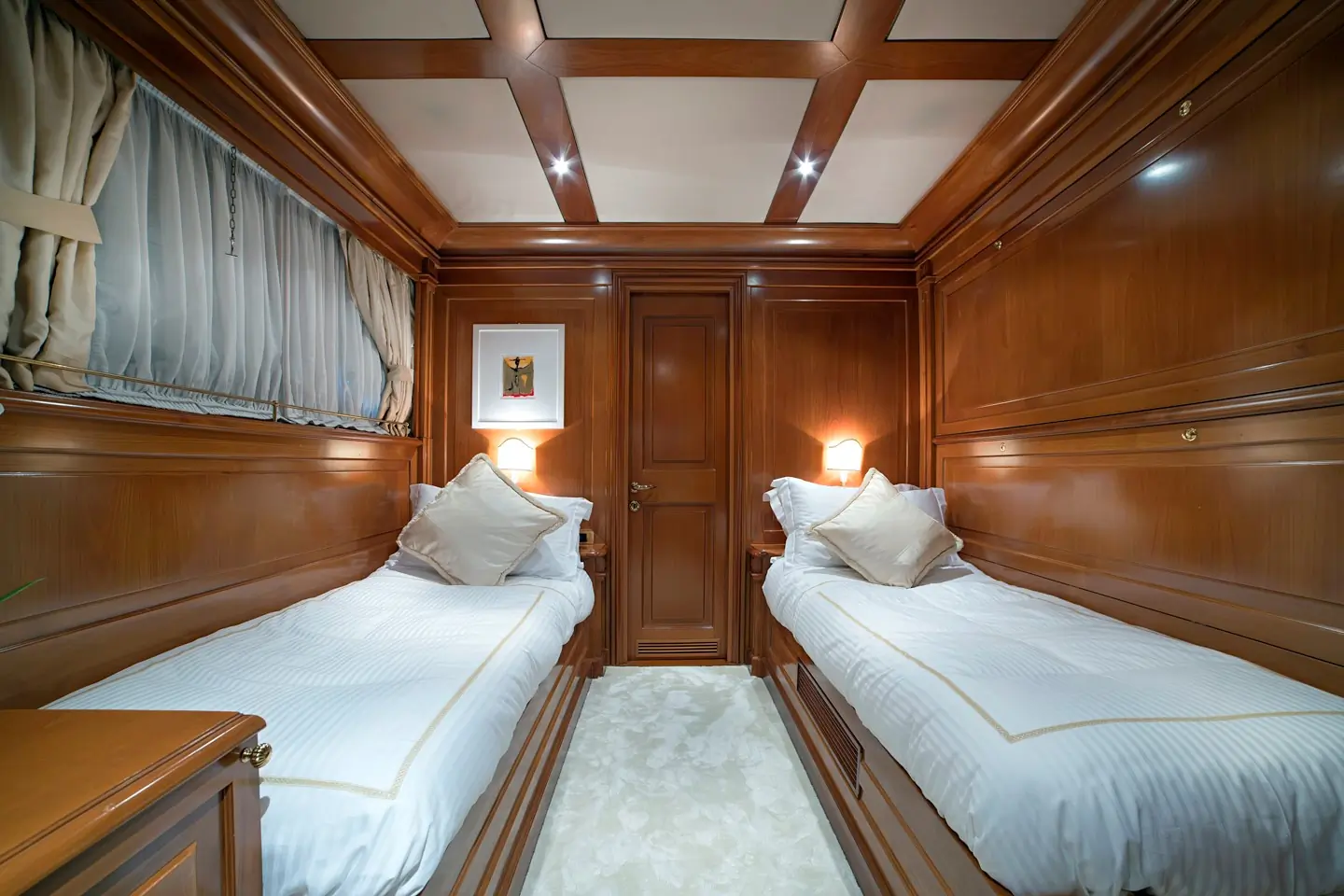Luxurious cabin with polished wood walls, two single beds with white linens, large pillows, soft lighting, a framed picture, and a curtained window. Ceiling beams and plush carpeting add elegance.