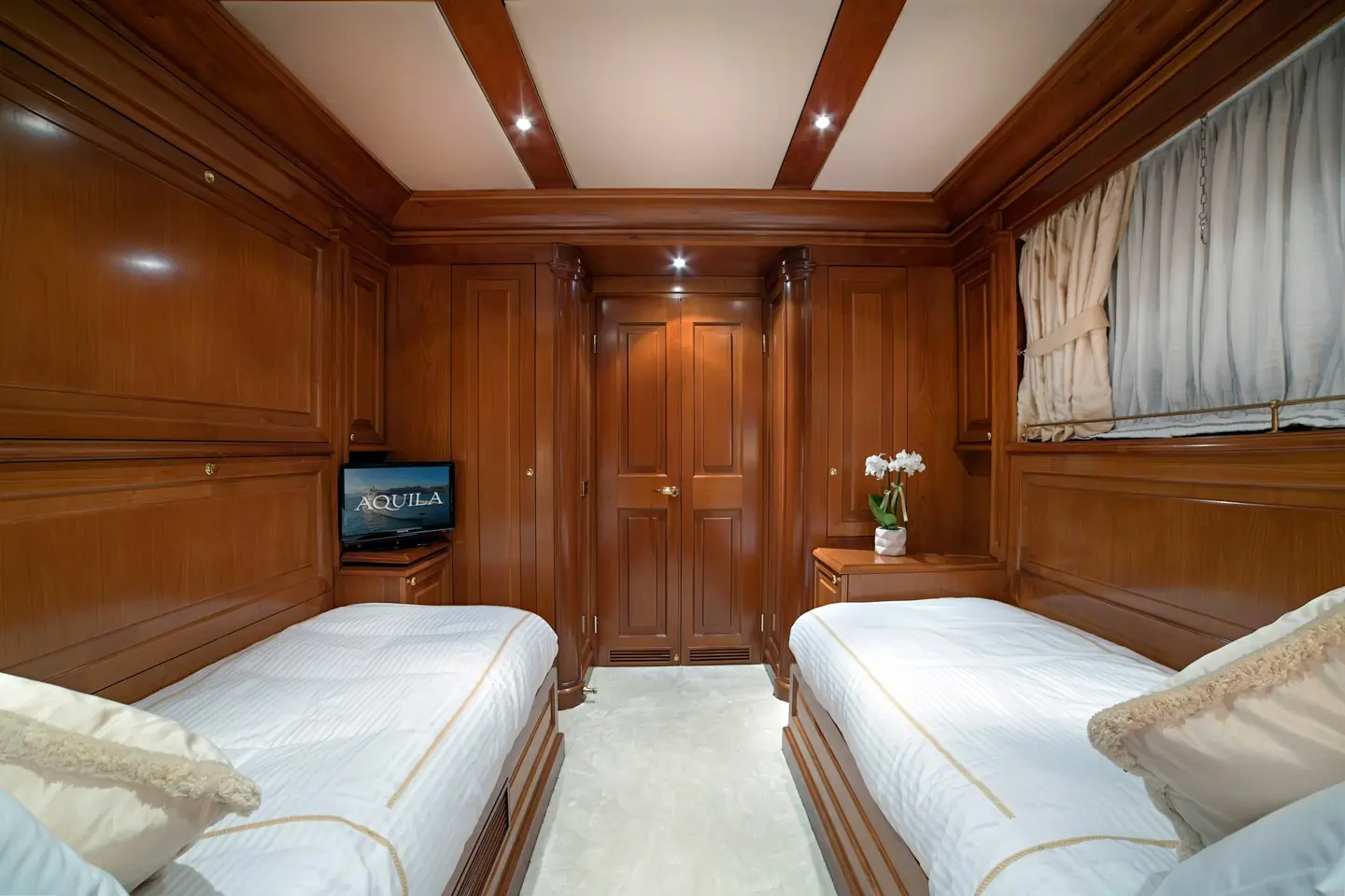A luxurious cabin with wooden walls, two single beds with white bedding, a small TV, a side table with flowers, and a curtained window. The lighting is warm and ceiling has wood accents.