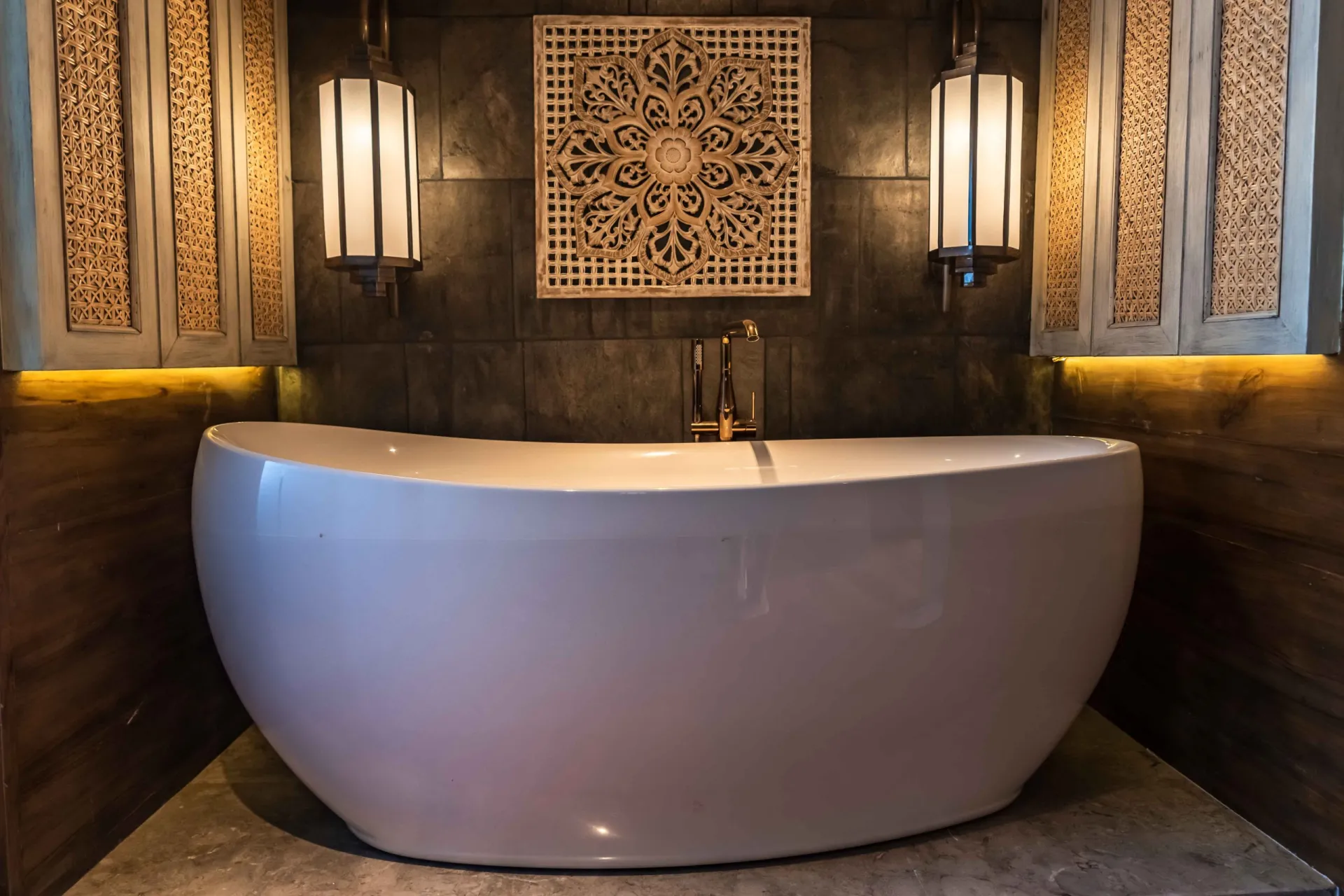 A modern freestanding white bathtub sits in a cozy alcove with wooden paneling, ornate wall art, and two large decorative hanging lanterns, creating a stylish and inviting bathroom space.