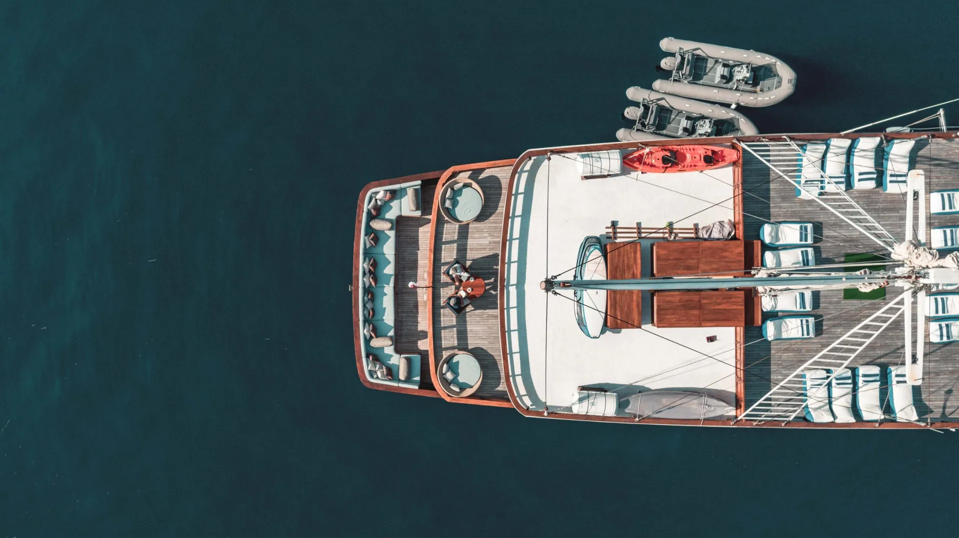 Aerial view of a yacht with loungers, seating areas, and small boats on deck, floating on calm, dark blue water. Two people are visible relaxing near the center of the yacht.
