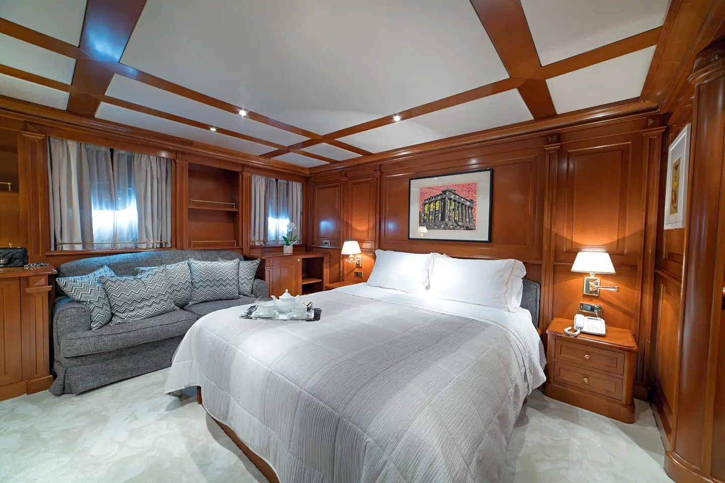 Luxurious bedroom with wood-paneled walls, a large bed with white bedding, a gray sofa, bedside tables with lamps, and a framed artwork above the bed.