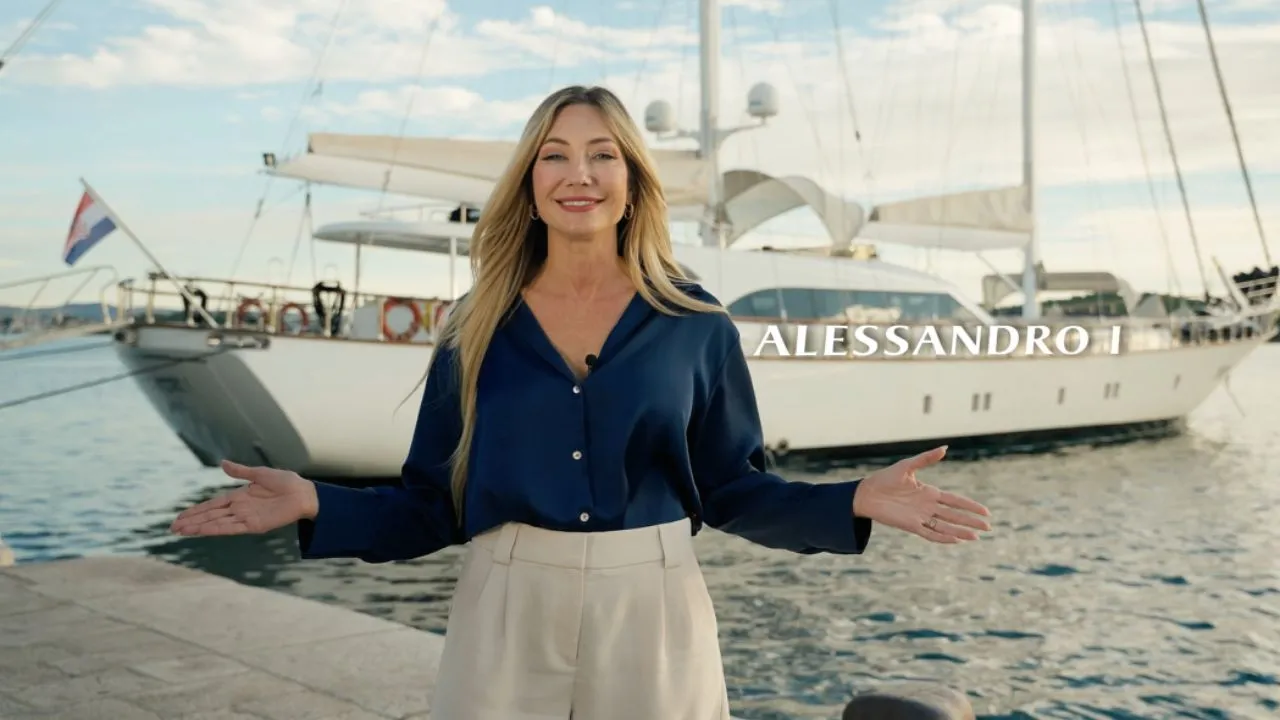 A smiling woman stands with open arms in front of a docked white yacht named ALESSANDRO I. The sky is clear and the water is calm, creating a welcoming and serene marina scene.