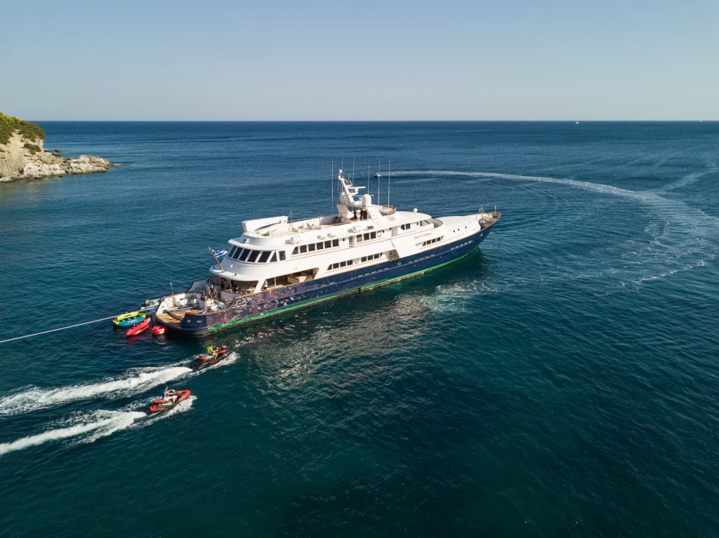 A large white and blue yacht is anchored in calm blue waters, with jet skis and small boats nearby. There is a forested coastline in the background under a clear sky.
