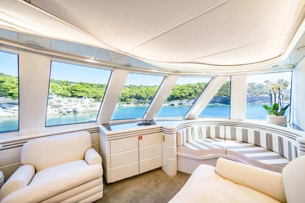 Interior of a modern yacht lounge with white furniture and large windows, offering a panoramic view of clear blue water and a green, tree-covered shoreline in the background. A potted orchid sits on a table by the window.
