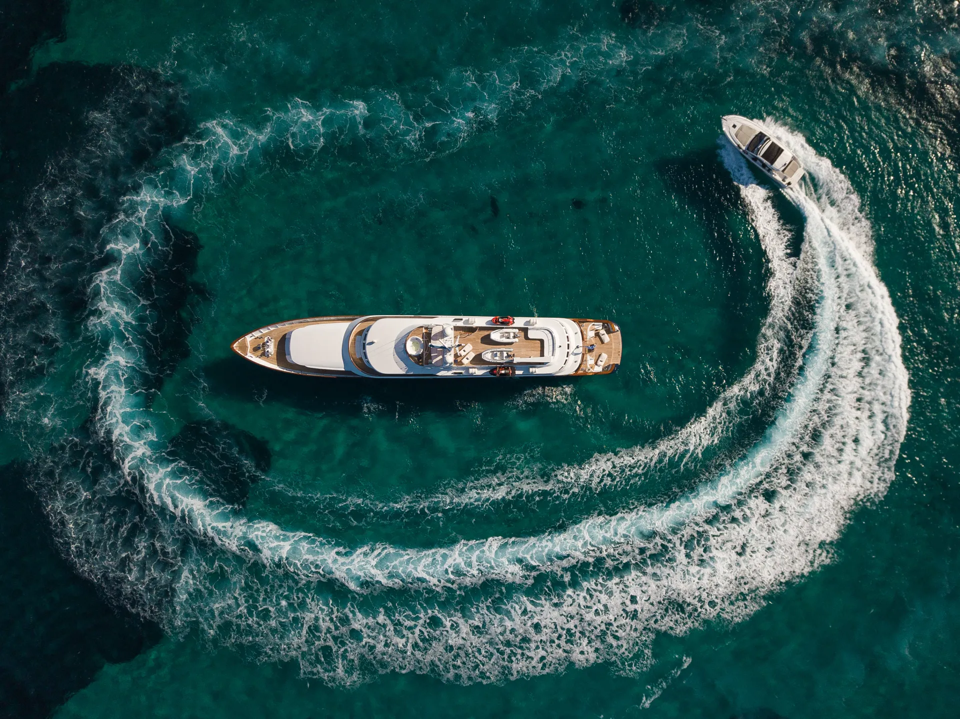 Aerial view of a large yacht on clear blue water, with a smaller boat circling it and creating a white wake in a spiral pattern.