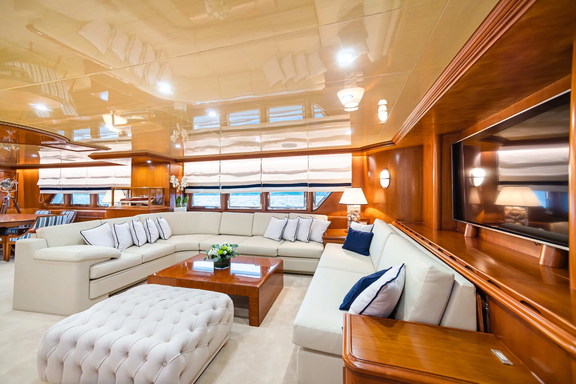 A luxurious yacht interior with cream-colored sectional sofas, a tufted ottoman, wooden tables, large windows with shades, and polished wood paneling reflecting warm light.