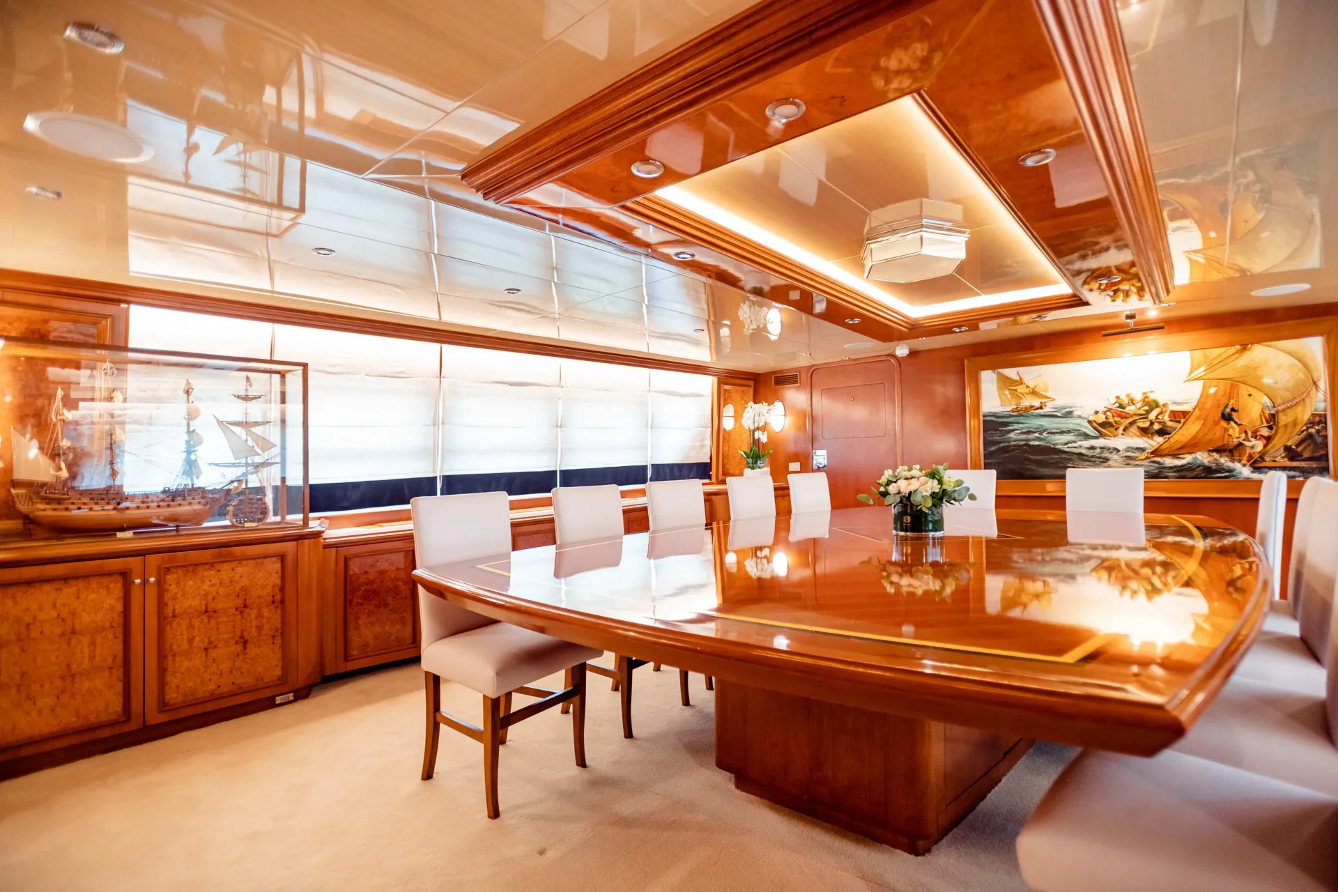 Luxurious yacht dining room with a polished wooden table, white cushioned chairs, model ship display, floral centerpiece, and nautical-themed artwork on the wall, under warm recessed lighting.