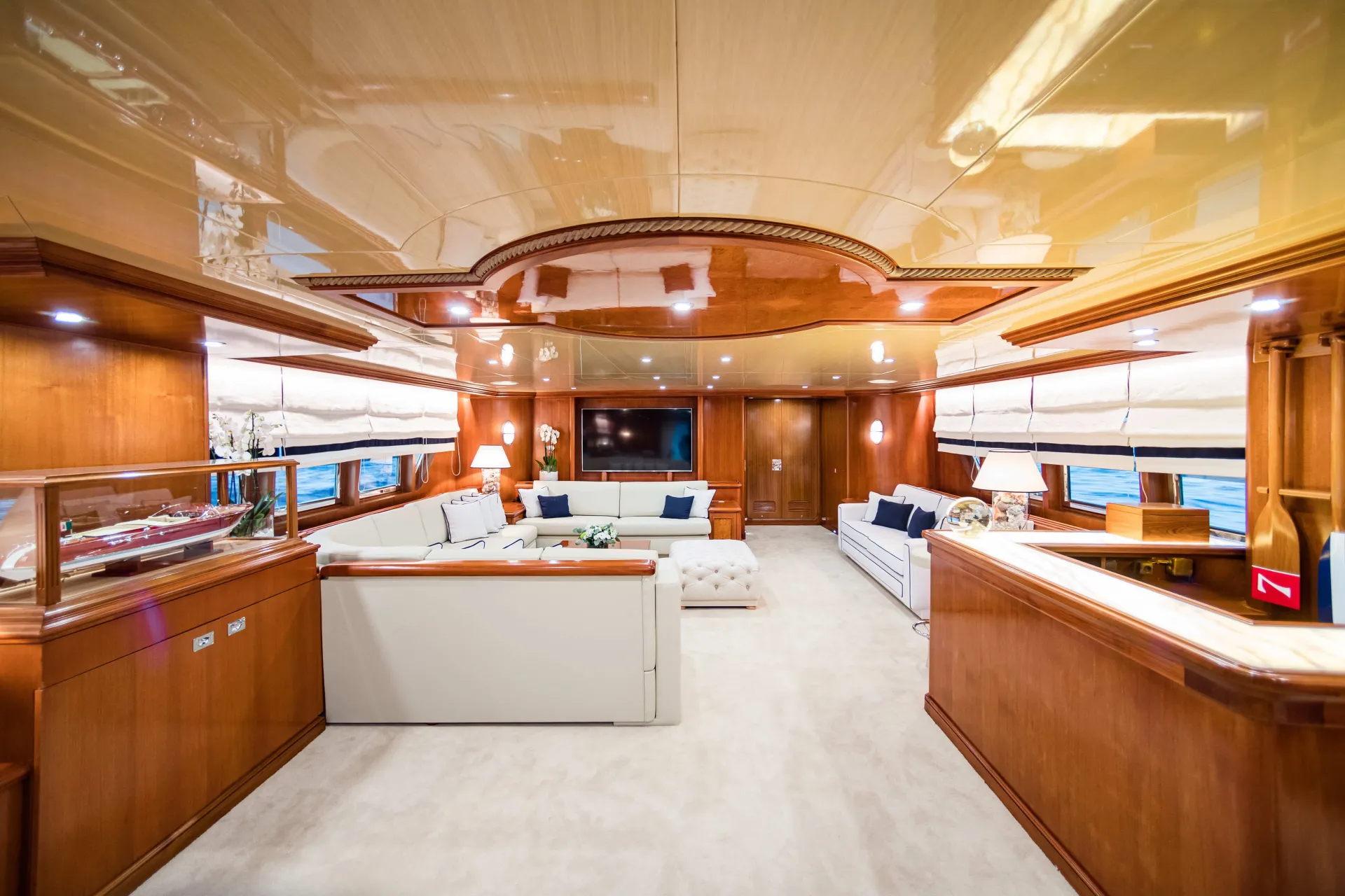 Spacious yacht interior with polished wood paneling, cream-colored carpet, white sofas, a flat-screen TV, large windows, and modern lighting, creating a luxurious and inviting lounge area.