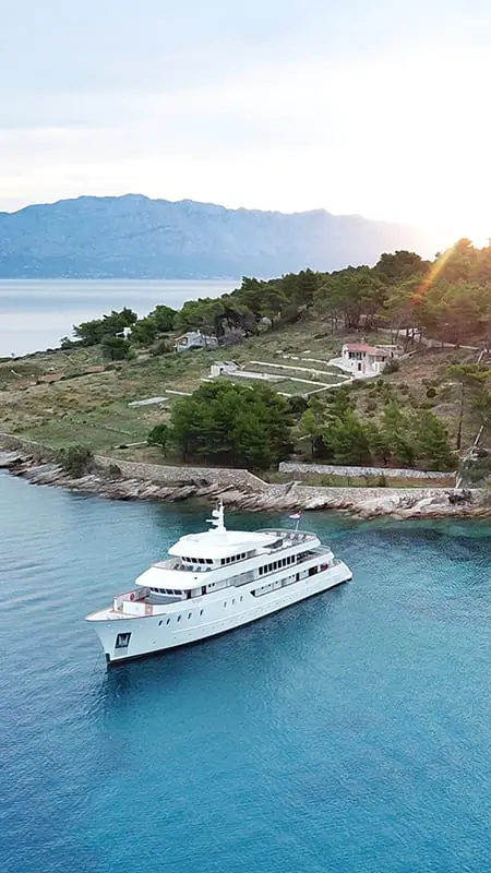 A white yacht sails near a rocky, tree-covered shoreline with a few houses. Clear blue water surrounds the boat and distant mountains are visible under a partly cloudy sky at sunset.