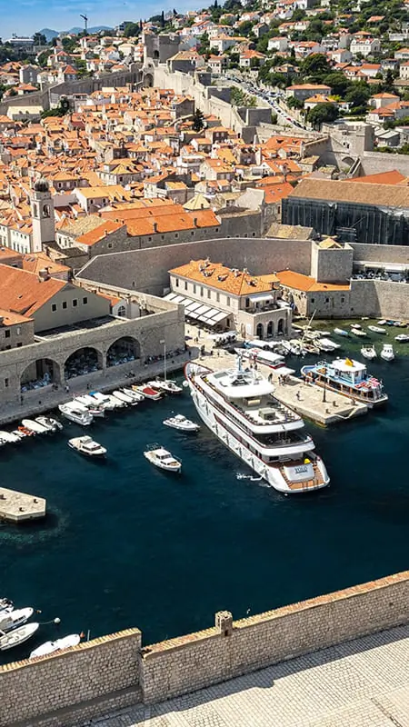 Aerial view of a historic coastal city with orange rooftops, stone walls, a harbor filled with boats, and a large yacht docked near the old town.