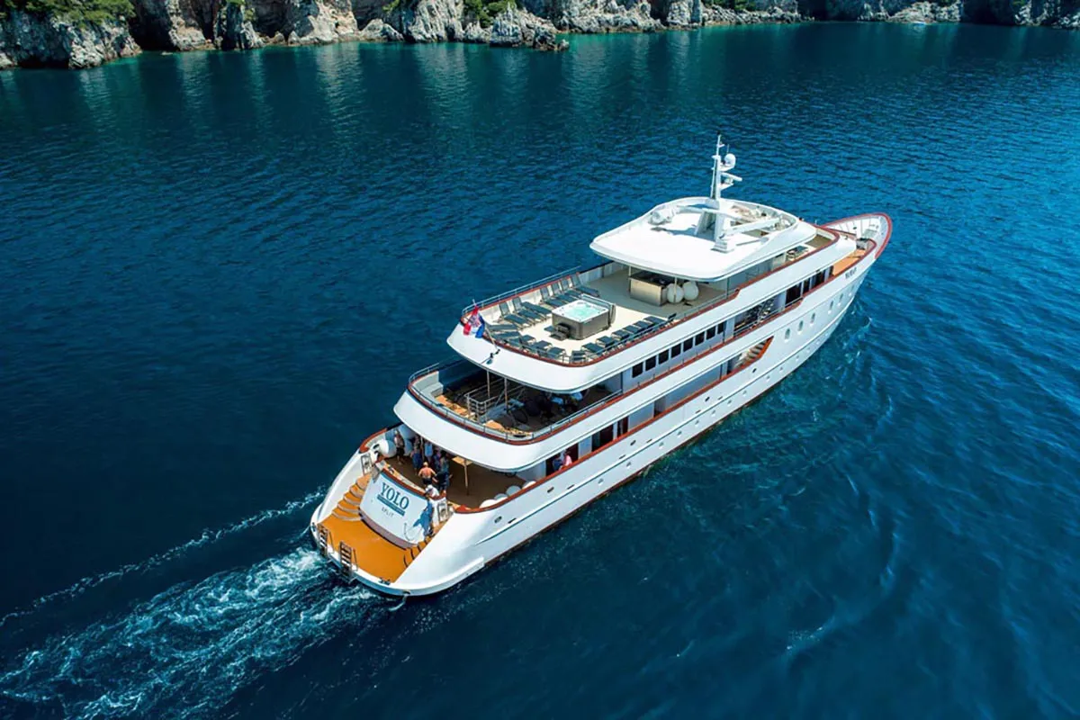 A large white yacht cruises through calm, deep blue water near a rocky, green shoreline under a clear sky. There are people relaxing on the upper deck.