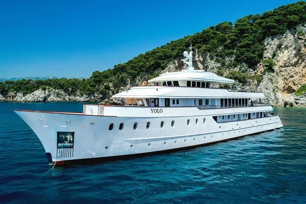 A large white luxury yacht named YOLO is anchored on calm blue water near a rocky, forested coastline under a clear blue sky.