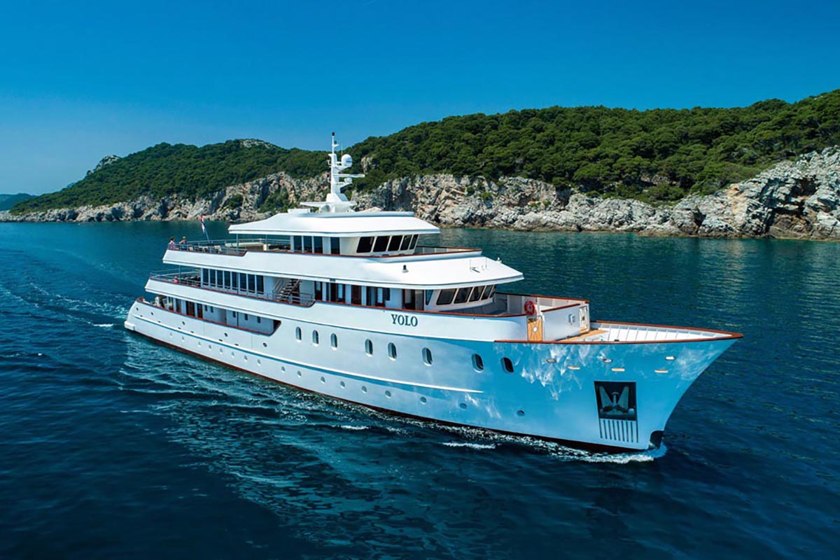 A large white luxury yacht named YOLO cruises on clear blue water near a rocky, forested coastline under a bright blue sky.