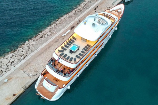 A large white yacht with a central pool and deck chairs is docked beside a stone pier, bordered by deep blue water and a rocky shoreline. People are gathered at the rear of the yacht.