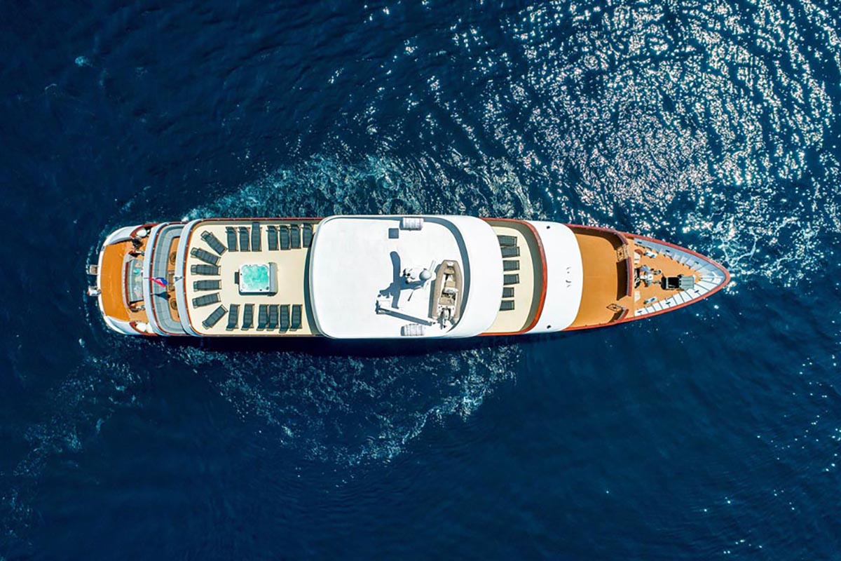 Aerial view of a large luxury yacht with a deck pool, sun loungers, and seating areas, cruising on deep blue water under clear skies.