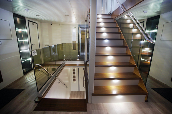 A modern indoor staircase with wooden steps and built-in step lights, glass railings, and a sleek, white interior design. The area is well-lit with recessed ceiling lights and features multiple doors and hallways.
