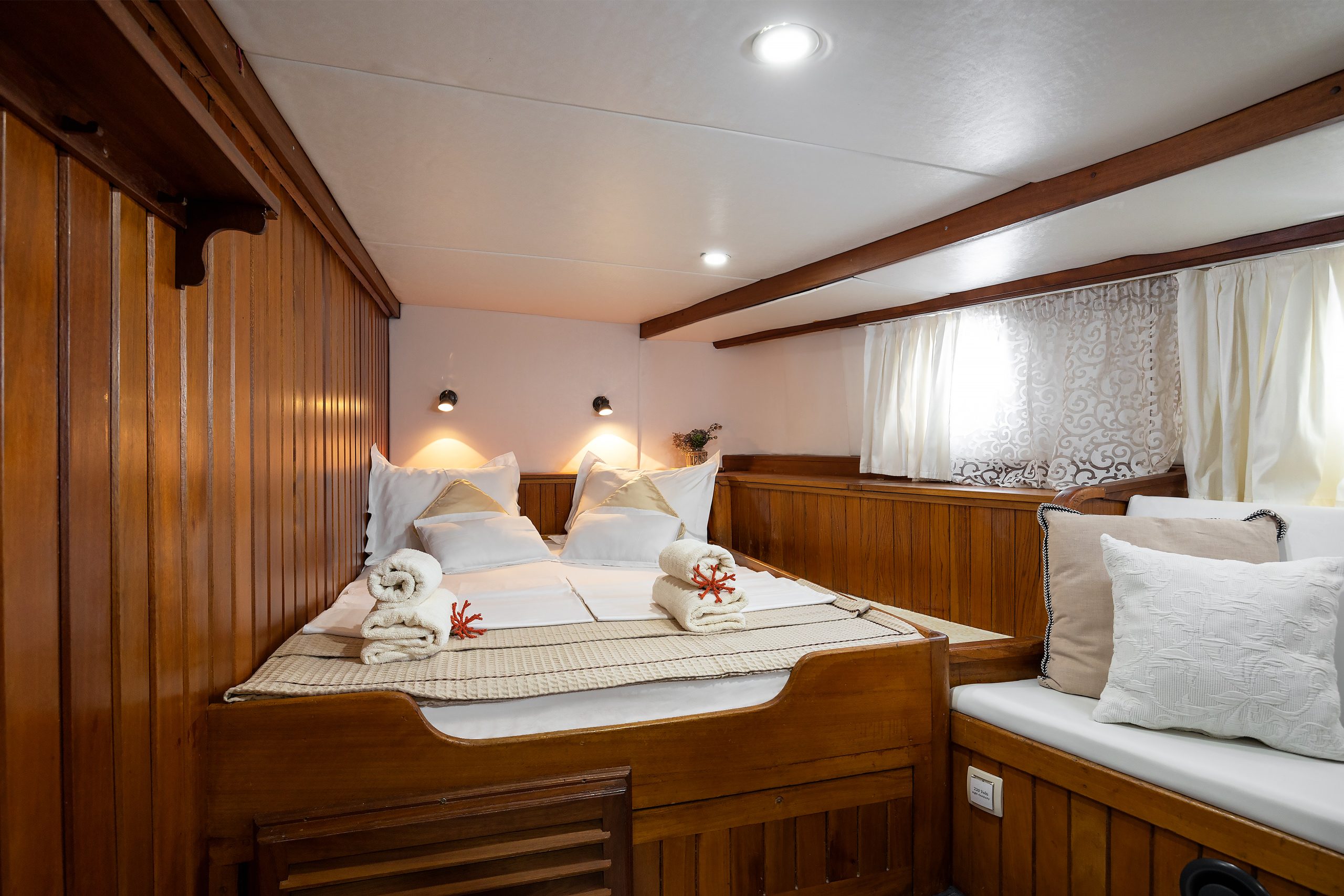 ABORDA A cozy wooden cabin bedroom features a neatly made double bed with white linens and towels, two reading lights above, wood-paneled walls, a small window with curtains, and a cushioned bench with pillows.