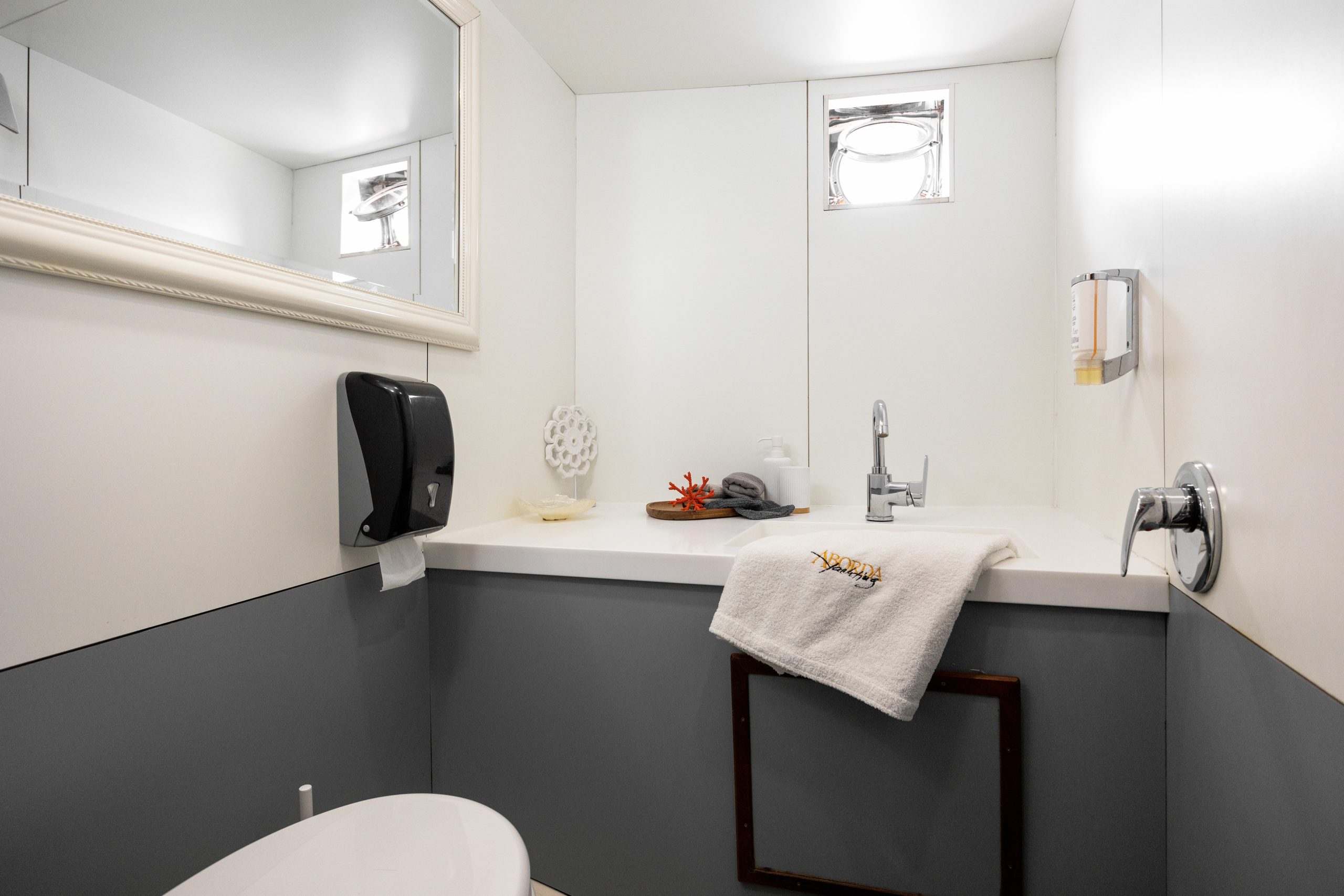 ABORDA A modern bathroom with white and gray walls, a mirror, a sink with a towel, a soap dispenser, a black paper towel holder, and a small window above the sink letting in light.
