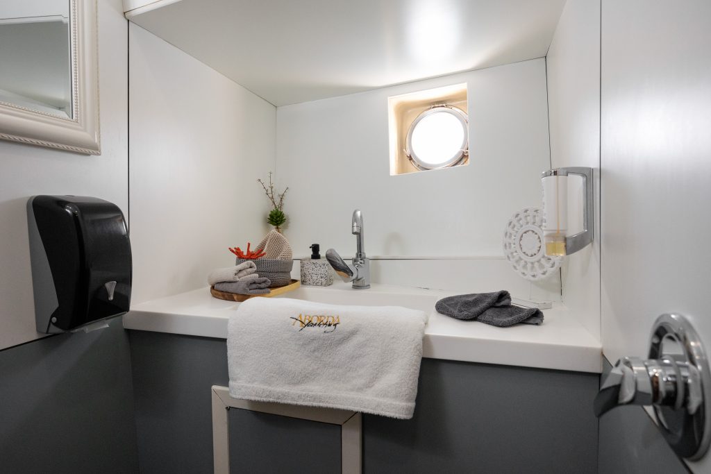 ABORDA A compact, modern bathroom features a white sink, a round porthole window, a towel embroidered with Nereids, folded grey towels, a soap dispenser, and small decorative items on the counter.