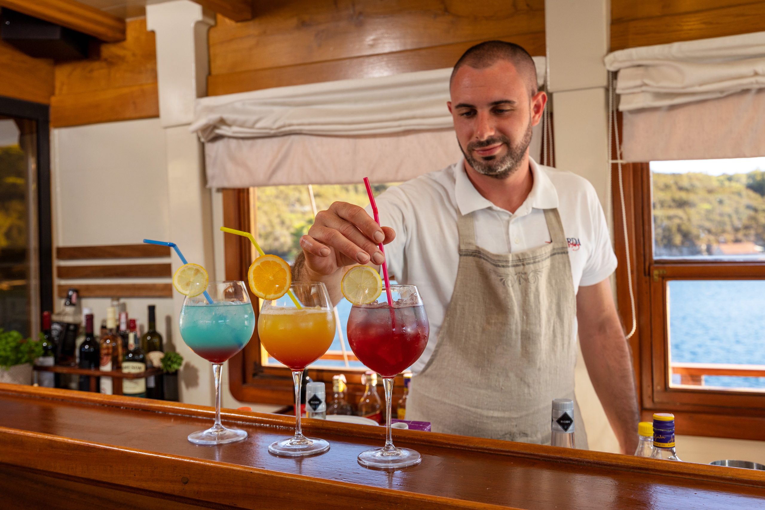 ABORDA A bartender places a straw in a red cocktail with a lemon slice garnish, next to two other colorful cocktails, behind a wooden bar with bottles in the background and sunlight streaming through windows.
