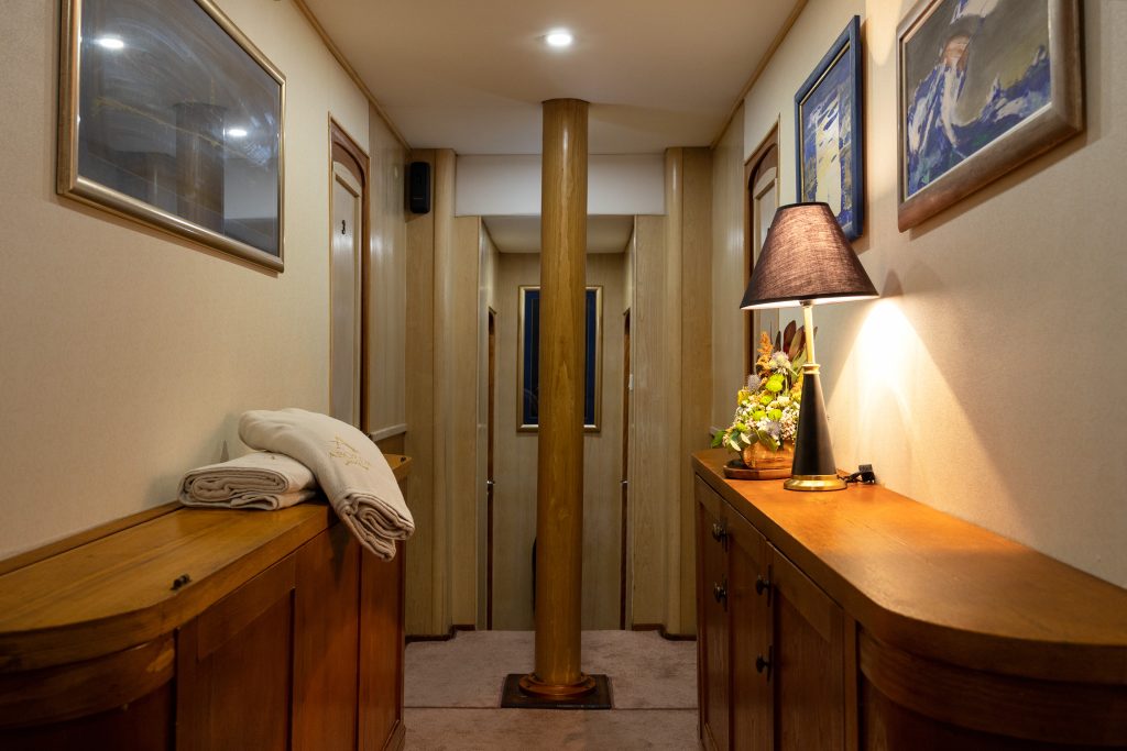 ABORDA A warmly lit hallway with wooden cabinets on both sides, a central column, art on the walls, a lamp, a small flower arrangement, and neatly folded towels resting on the left cabinet.