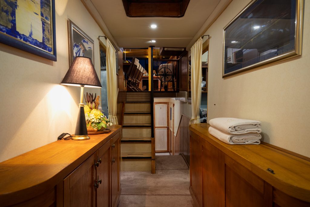 ABORDA A cozy hallway inside a boat or yacht with wooden cabinets, a lamp, framed pictures, folded towels, and stairs leading up to another area with visible chairs and tables.