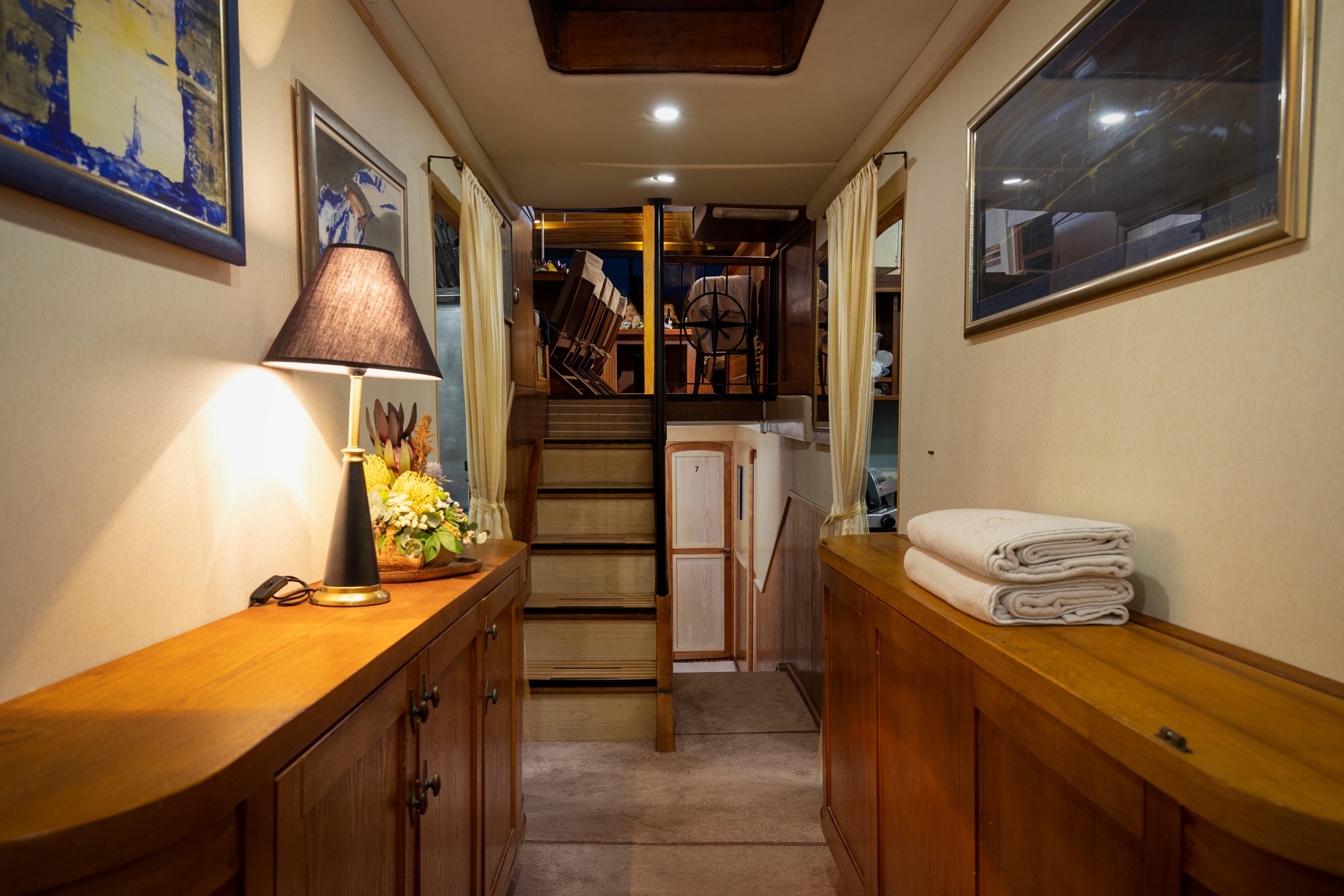 ABORDA A cozy hallway inside a boat or yacht with wooden cabinets, a lamp, framed pictures, folded towels, and stairs leading up to another area with visible chairs and tables.
