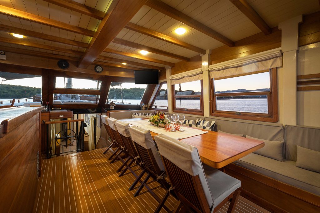 ABORDA A cozy yacht dining area with a wooden table set for six, cushioned benches, wooden chairs, large windows showing a waterfront view, and warm ceiling lights illuminating the space.