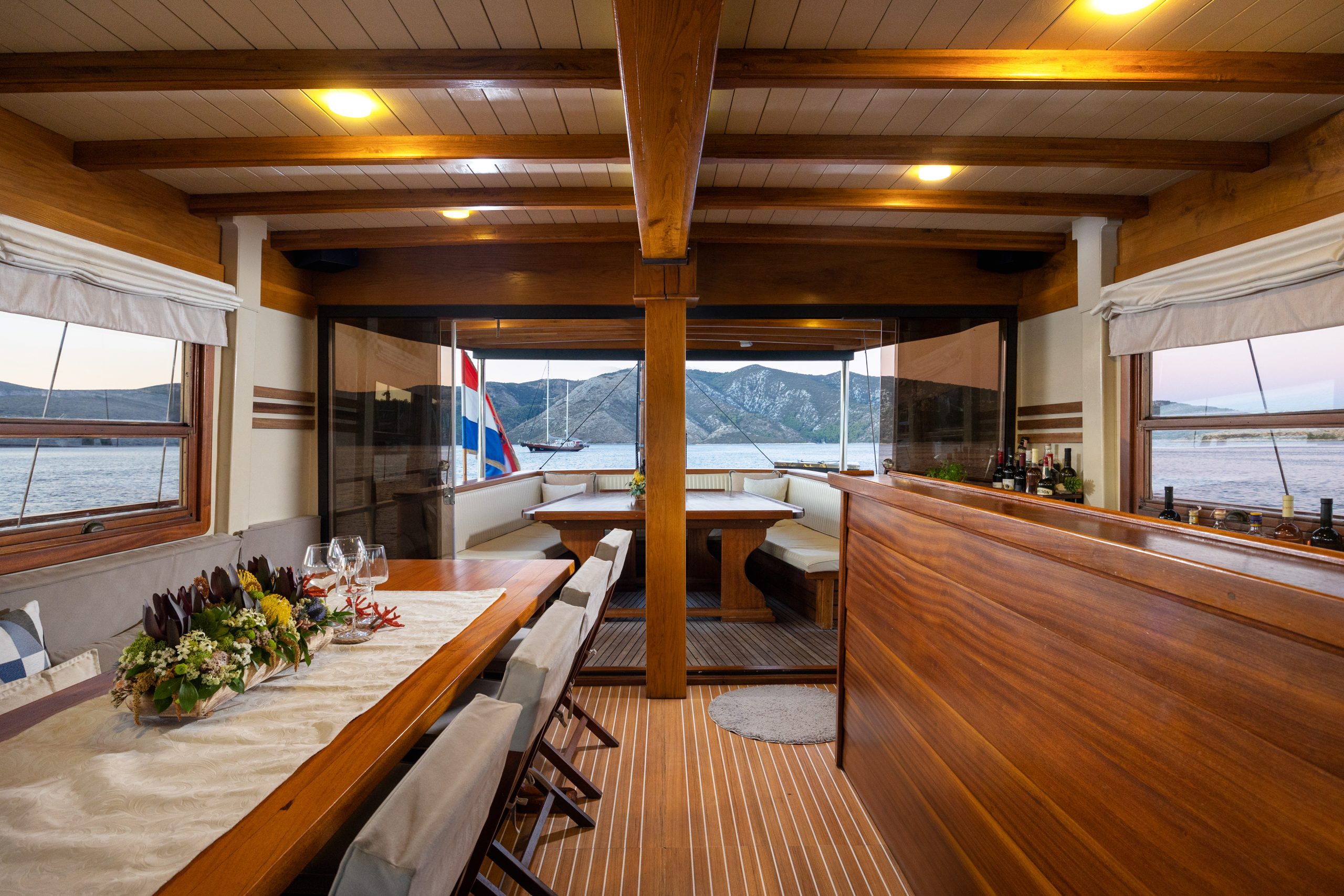 ABORDA Interior of a wooden yacht dining area set with a decorated table, chairs, and large windows showing views of water and distant mountains at sunset. A flag is visible outside the back window.