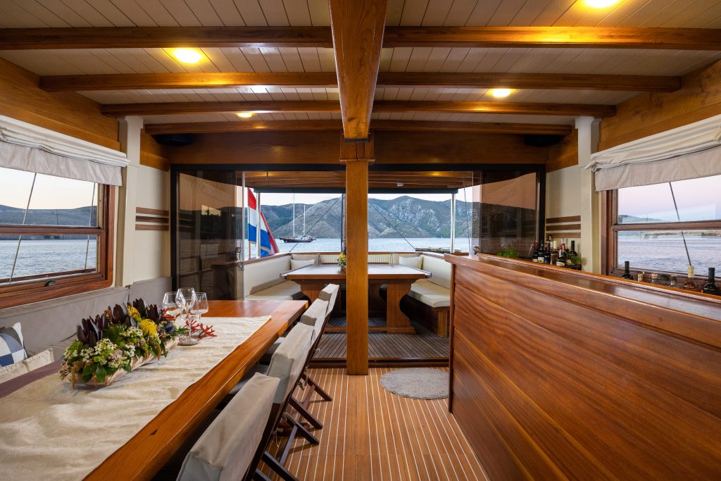 ABORDA Elegant yacht interior with a wooden dining table set for a meal, surrounded by chairs. Large windows reveal a scenic view of mountains and water, with evening light streaming inside.