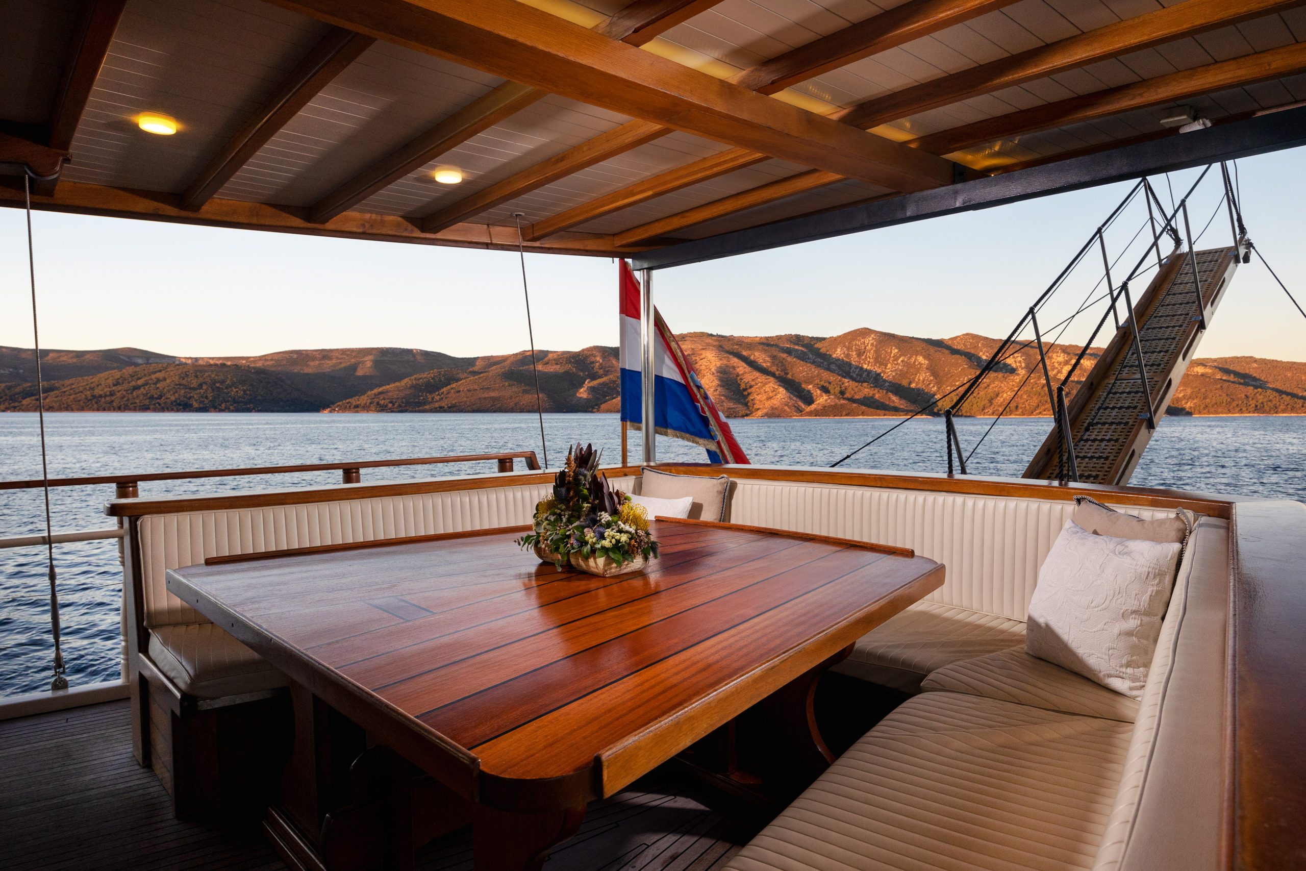 ABORDA A wooden table with a flower arrangement sits on a yachts covered deck, surrounded by cushioned benches. A Dutch flag is visible at the stern, with calm water and distant hills at sunset in the background.