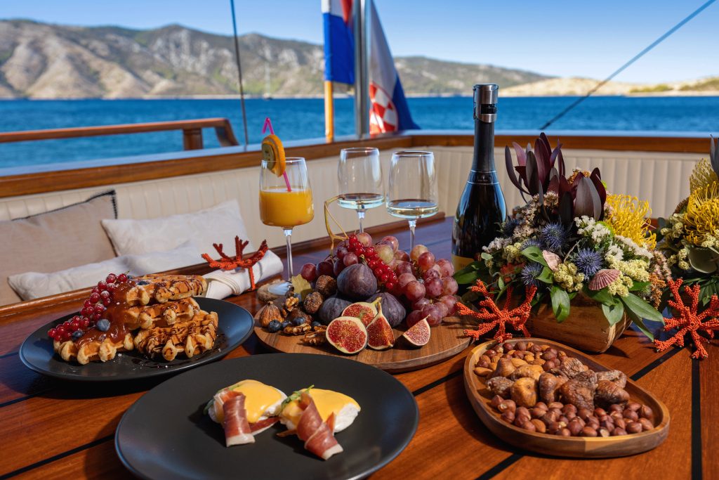 ABORDA A wooden table on a boat is set with plates of waffles, figs, grapes, prosciutto with eggs, mixed nuts, a bottle of wine, glasses, a flower arrangement, and ocean and mountains in the background.