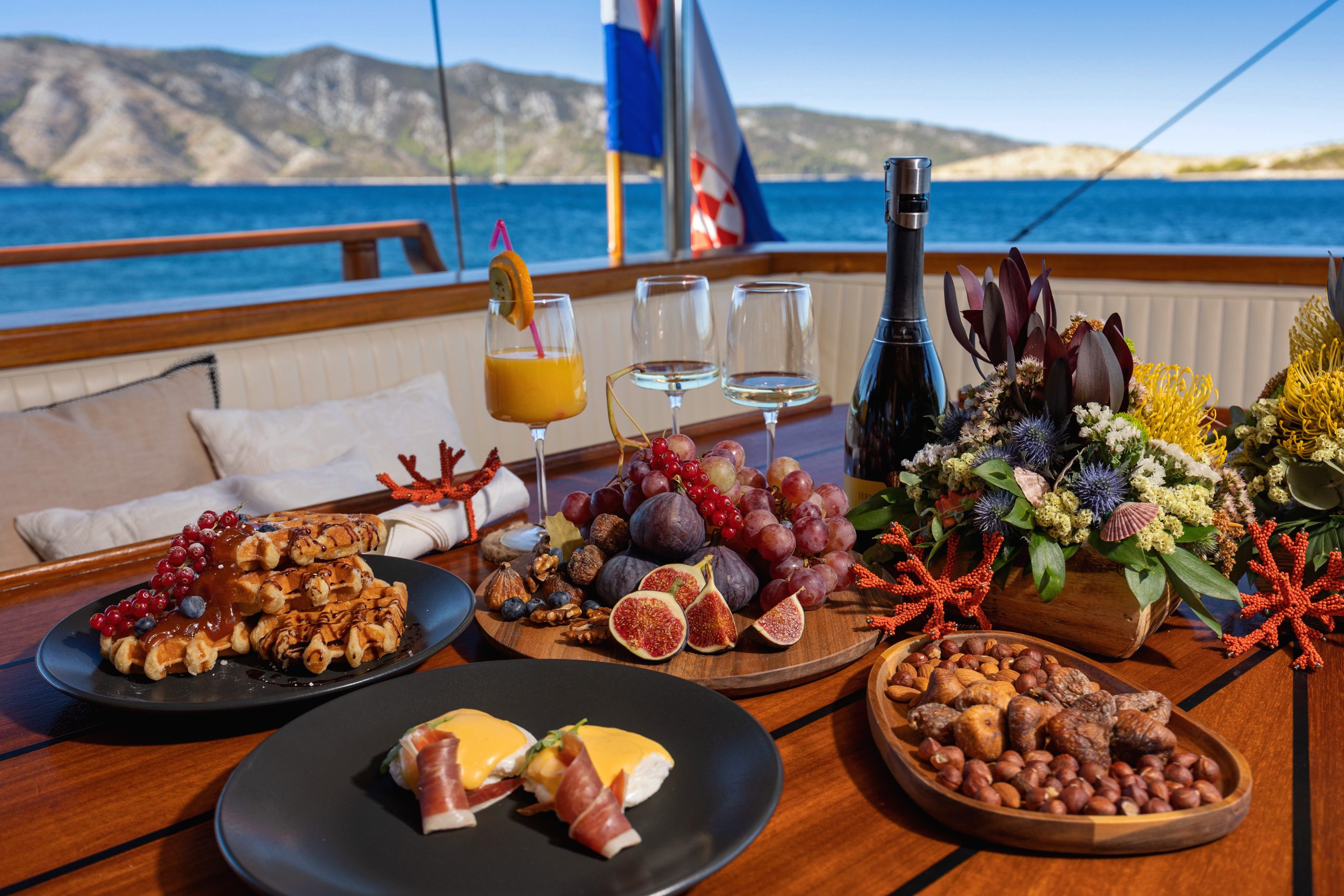 ABORDA A wooden table on a boat is set with plates of waffles, figs, grapes, prosciutto with eggs, mixed nuts, a bottle of wine, glasses, a flower arrangement, and ocean and mountains in the background.