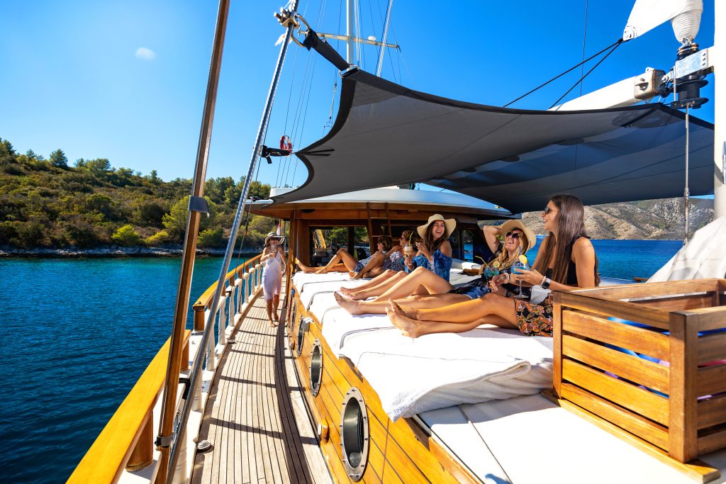 ABORDA A group of people relax on sun loungers under a shade on the deck of a yacht, enjoying drinks and the sunshine, with blue water and green hills in the background.