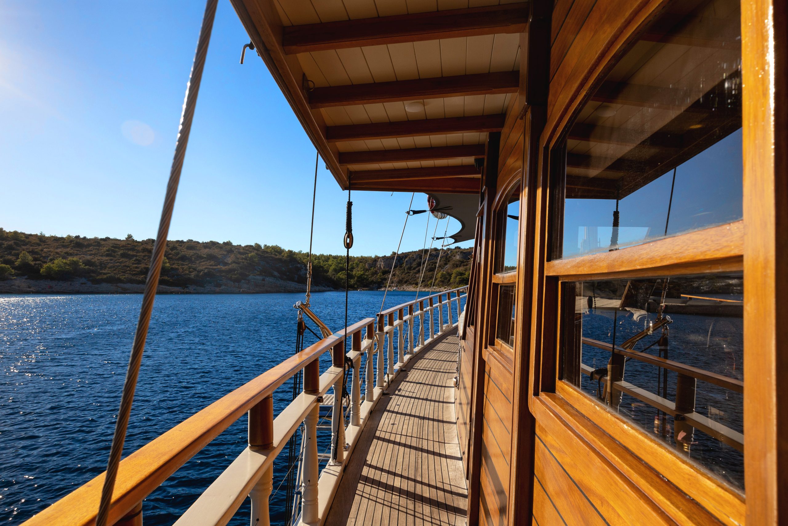 ABORDA View from the side deck of a wooden boat sailing near a coastline, with clear blue water, a sunny sky, and green hills in the distance.